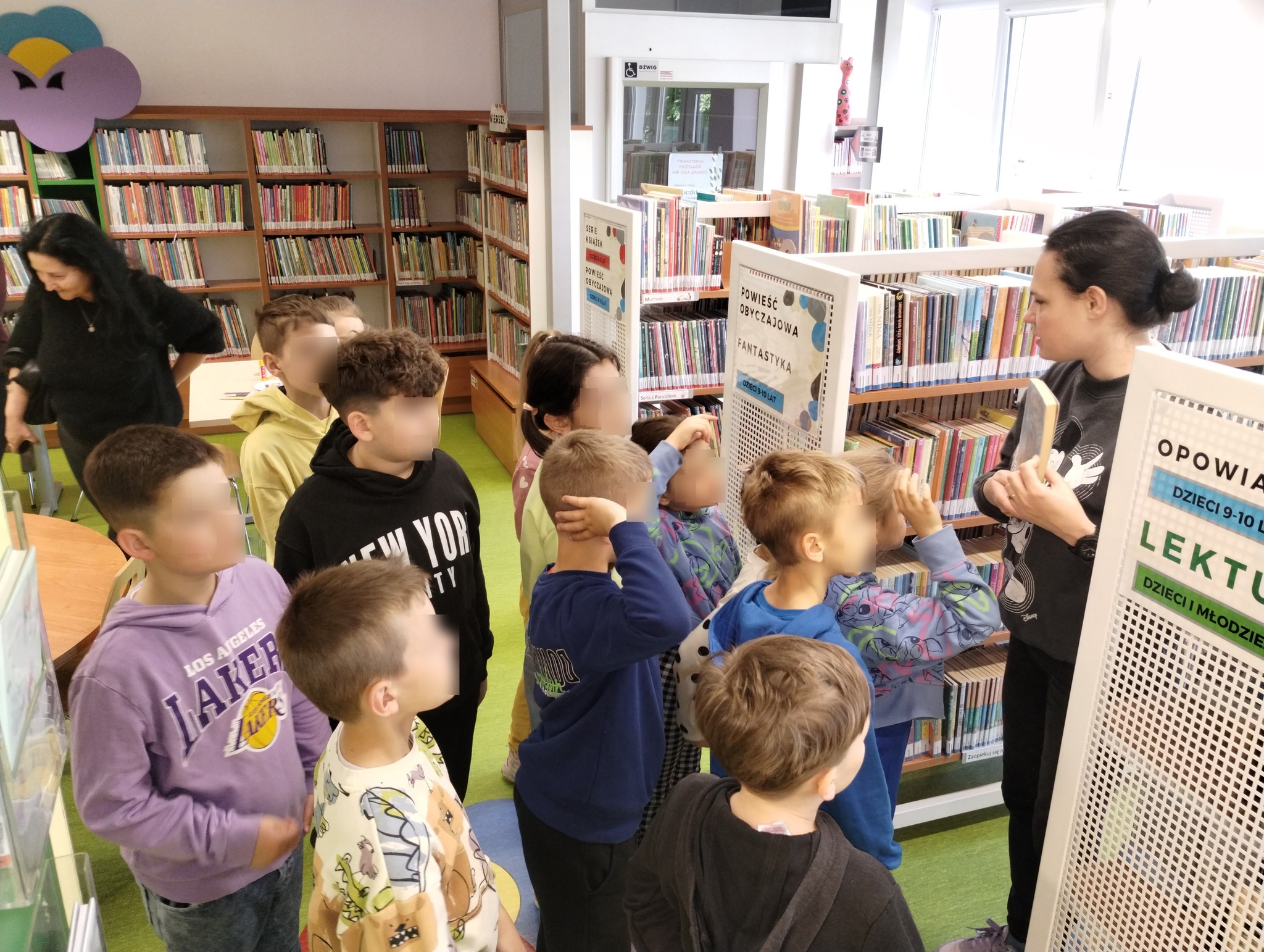 grupa dzieci stoi przed regałami z książkami, przed nimi stoi kobieta