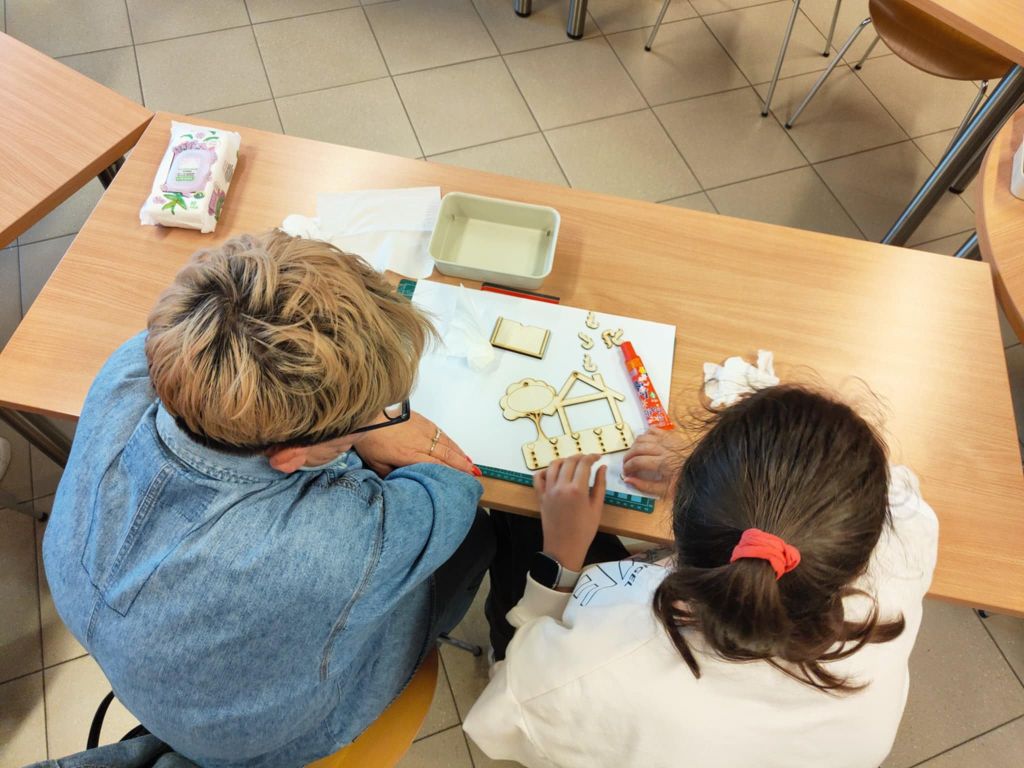 Na zdjęciu widoczne są dwie osoby przy stole, pracujące nad drewnianymi wieszakami na klucze. Na blacie znajdują się różne materiały i narzędzia, w tym linijka, klej w sztyfcie oraz haczyki do zawieszenia kluczy. W tle widać inne stoły i krzesła, co sugeruje większą przestrzeń warsztatową.