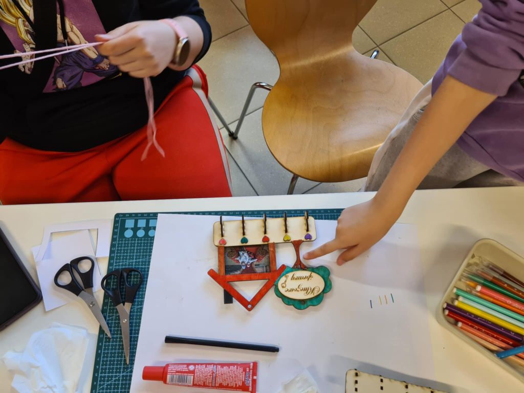 Dwie osoby pracują nad rękodziełem przy stole. Na blacie znajdują się nożyczki, markery, klej, papier i mata do cięcia. Tworzony przedmiot to drewniana ramka ze zdjęciem, ozdobnymi elementami i kolorowymi koralikami na sznurkach.