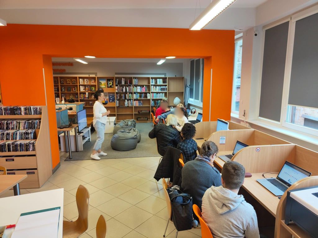 Po prawej stronie widać osoby dorosłe oraz młodzież siedzące przy laptopach w sali w bibliotece. Za ich plecami stoi kobieta prowadząca zajęcia.