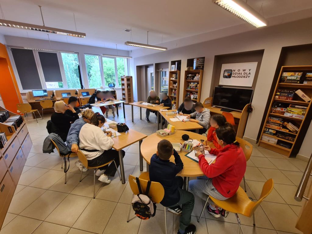 Zdjęcie przedstawia otwartą przestrzeń biblioteki, gdzie grupa osób bierze udział w warsztatach siedząc przy stołach. Widać regały, stanowiska komputerowe oraz miejsca do pracy.
