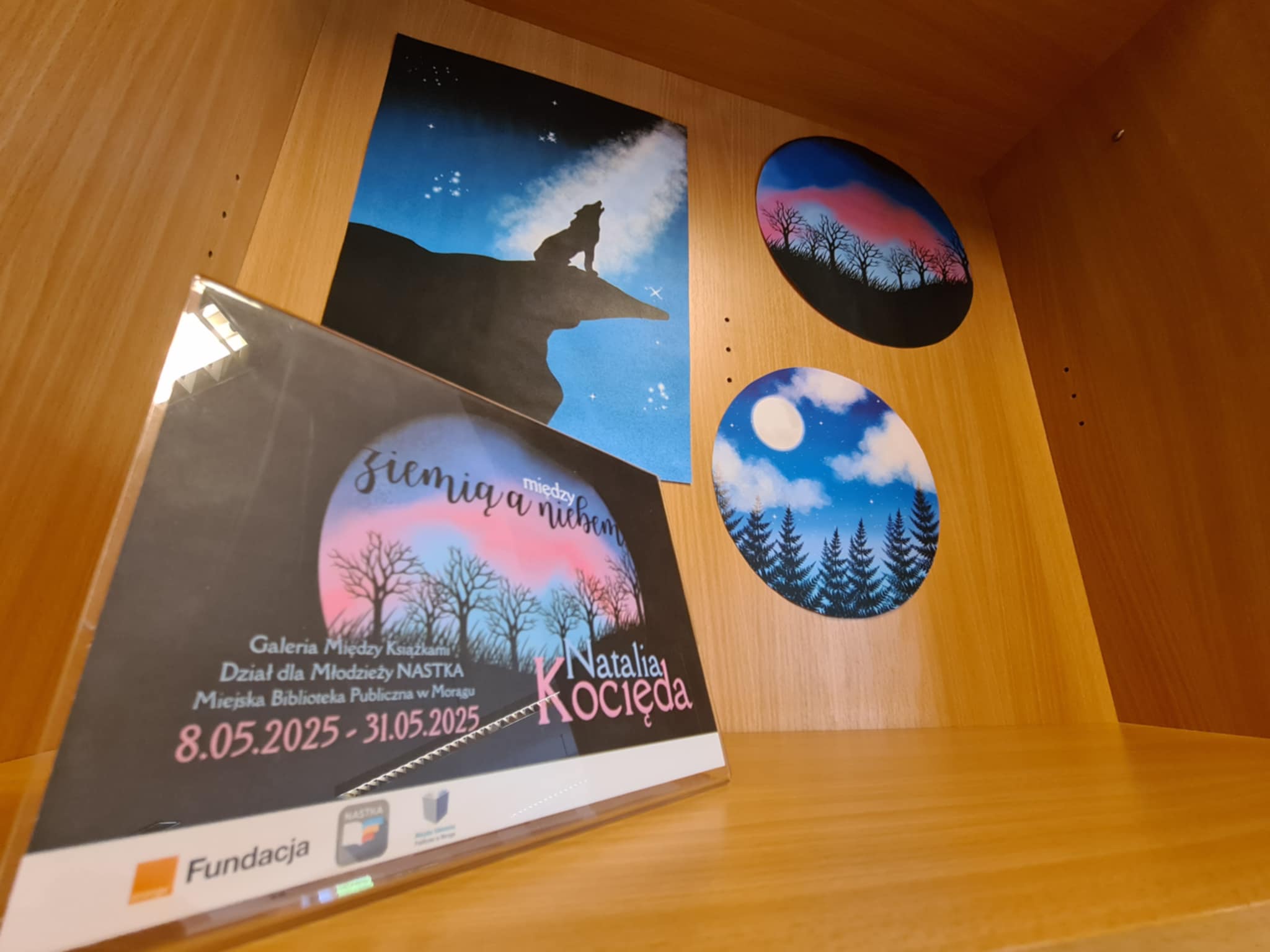 Wystawa sztuki w bibliotece – obrazy nocnych krajobrazów na drewnianej półce.