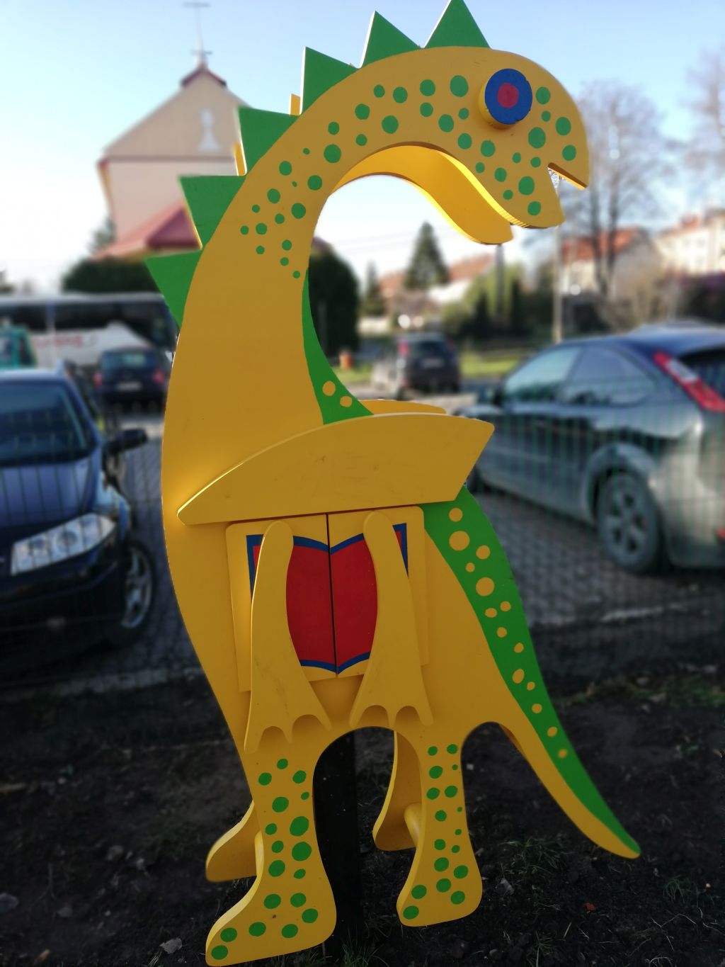 Kolorowa mikrobiblioteka w kształcie żółtego dinozaura z zielonymi kropkami i grzbietem z zielonych trójkątów. Dinozaur stoi na tle zaparkowanych samochodów i budynku. Mikrobiblioteka ma zamykane drzwiczki w kształcie otwartej książki, z czerwonym wnętrzem i niebieskim konturem. W dolnej części widoczne są żółte łapy dinozaura. Całość ustawiona jest na metalowym słupku, wbitym w ziemię.