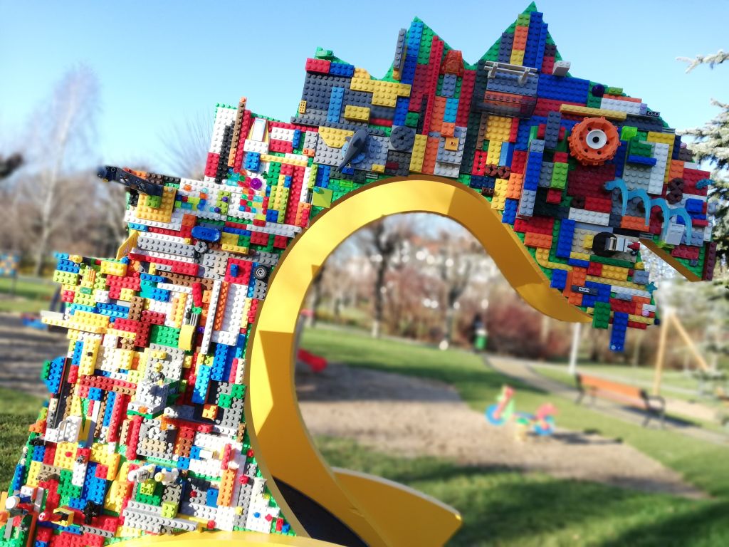 Cześć mikrobiblioteki przedstawiająca głowę smoka ozdobionego klockami lego. Głowa smoka jest zamocowana na żółtym, zakrzywionym elemencie, który tworzy jego szyję. W tle widać zieloną trawę, drzewa oraz plac zabaw z piaskownicą i huśtawką.