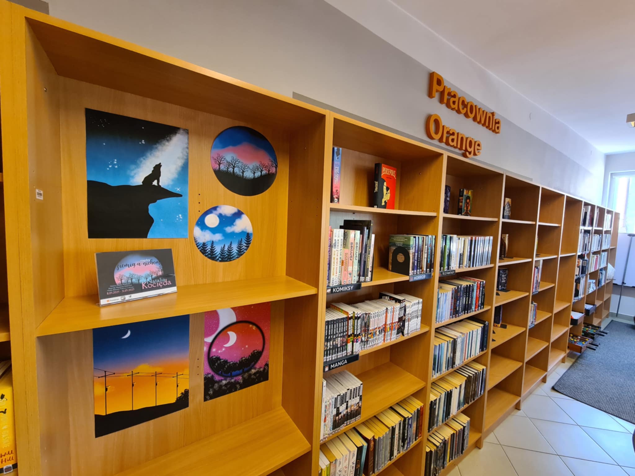 Biblioteka z drewnianymi półkami pełnymi książek oraz wystawą obrazów nocnych krajobrazów.