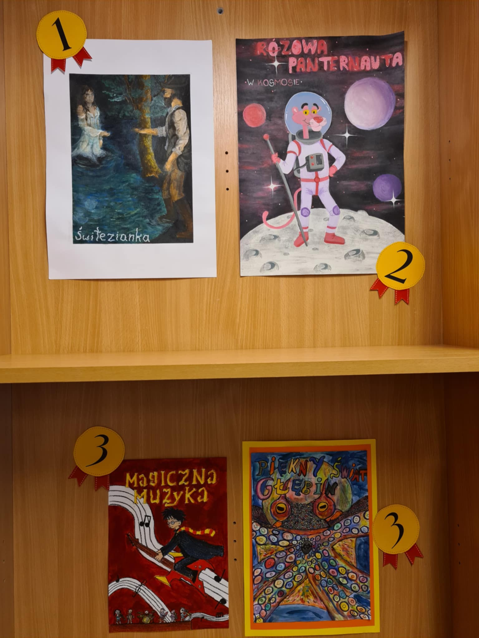 Drewniana półka z czterema nagrodzonymi ilustracjami konkursowymi. Na górze „Świtezianka” (miejsce 1) i „Różowa Panternauta” (miejsce 2). Na dole dwie ilustracje ex aequo na miejscu 3: „Magiczna muzyka” — postać przypominająca Harry'ego Pottera lecąca na gitarze po pięciolinii oraz „Piękny świat głębin” — kolorowy ośmiornica na tle podwodnego świata.