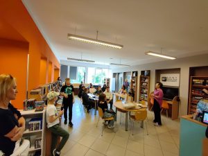 Zdjęcie przedstawia rozdanie nagród podczas konkursu plastycznego w bibliotece. W sali grupa młodzieży oraz dorosłych siedzi przy stole i stoi wokół. Po prawej stronie stoi kobieta prowadząca spotkanie.