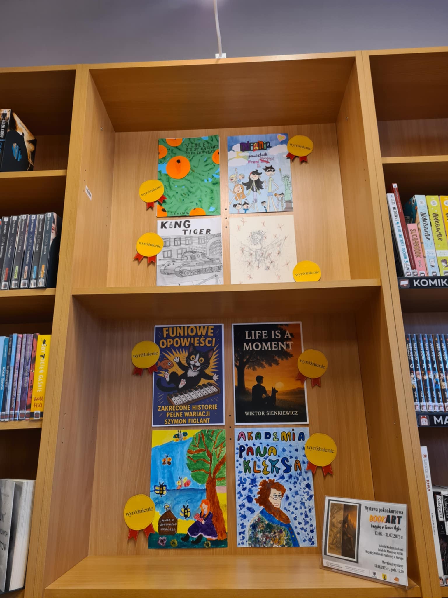 Drewniana biblioteczna półka z ośmioma wyróżnionymi pracami konkursowymi. Na górze m.in. ilustracja z dyniami, rysunek dzieci z napisem „Niania”, szkic czołgu „King Tiger” i abstrakcyjny rysunek ołówkiem. Na dole plakaty książkowe: „Funiowe opowieści”, „Life is a moment”, „Akademia Pana Kleksa” i praca z osobą czytającą nad jeziorem. Każda praca oznaczona żółtym medalem z napisem „wyróżnienie”.