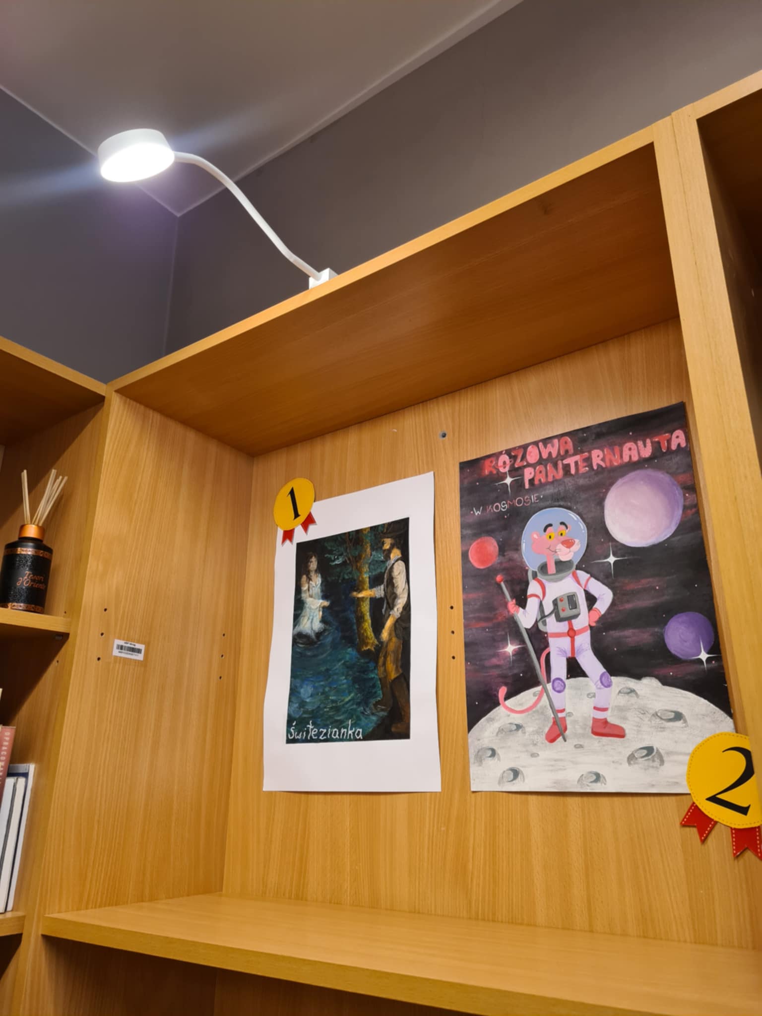 Drewniana półka biblioteczna z dwiema ilustracjami konkursowymi. Po lewej stronie ilustracja „Świtezianka” przedstawiająca kobietę w wodzie i mężczyznę na brzegu, stylizowana na obraz malarski. Po prawej ilustracja „Różowa Panternauta w kosmosie” — postać Różowej Pantery w stroju astronauty na powierzchni Księżyca, z kolorowymi planetami w tle. Nad ilustracjami numery miejsc: pierwsze i drugie.