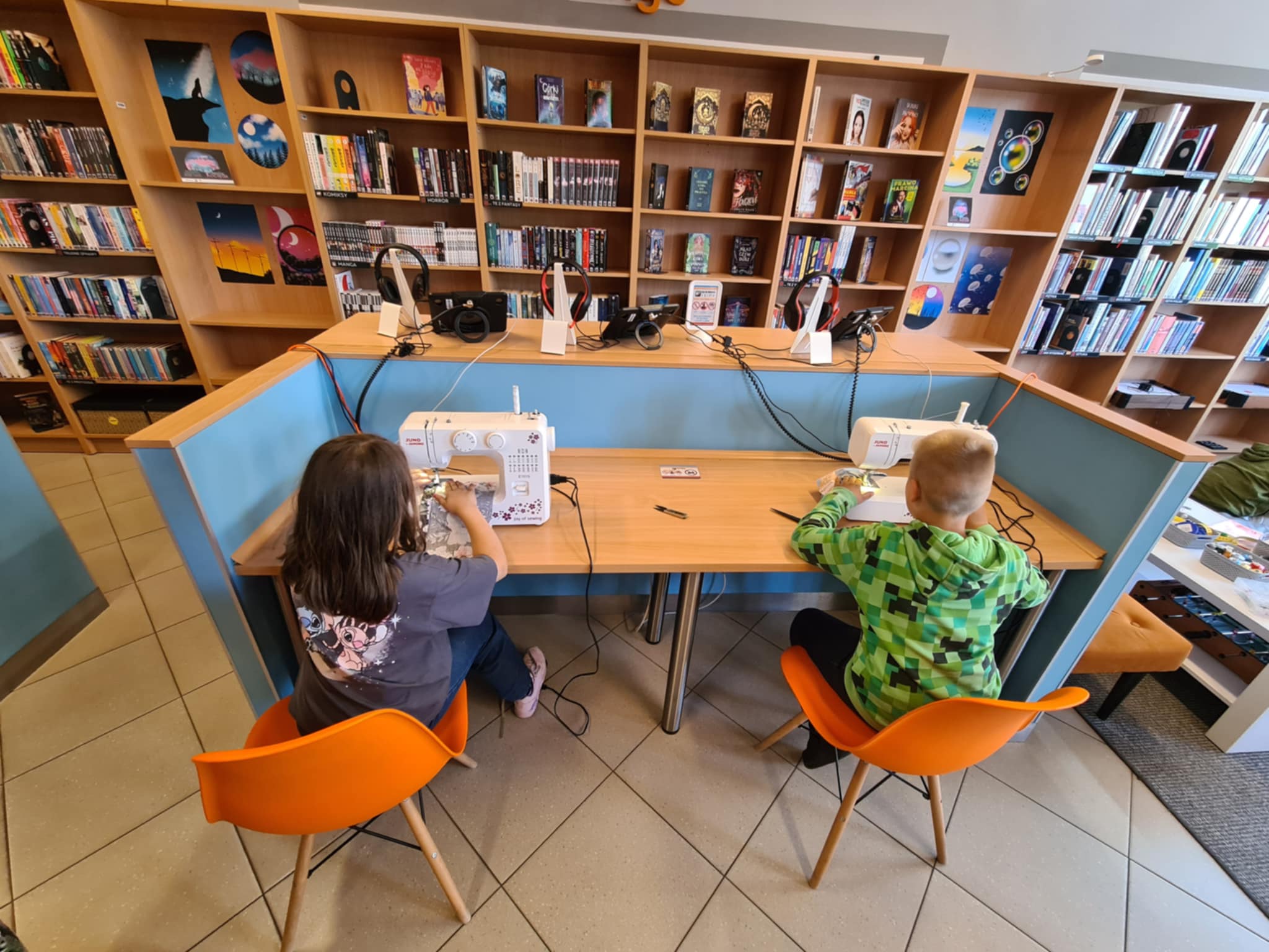 Dwoje dzieci siedzi przy maszynach do szycia i szyje w bibliotece podczas warsztatów krawieckich.