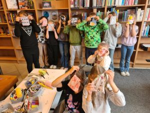 Grupa dzieci w bibliotece trzyma przed twarzami własnoręcznie uszyte woreczki podczas warsztatów krawieckich.