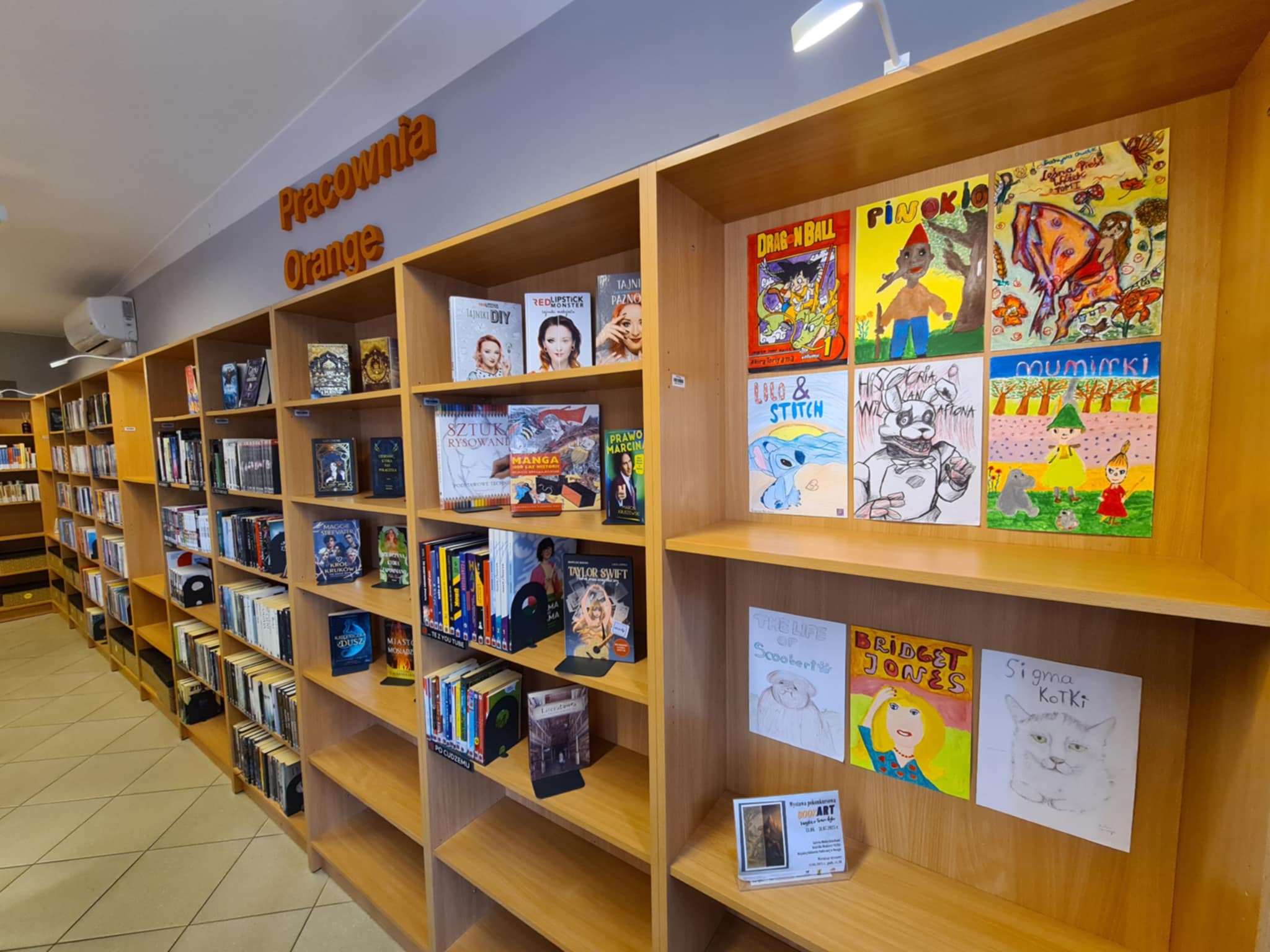 Szeroki widok na przestrzeń biblioteki Pracowni Orange z regałami pełnymi książek. Na przedniej półce widoczne prace konkursowe inspirowane popkulturą: „Dragon Ball”, „Pinokio”, „Lilo i Stitch”, „Hello Kitty”, „Muminki”, „The life of Scooby”, „Bridget Jones” i „Sigma Kotki”.
