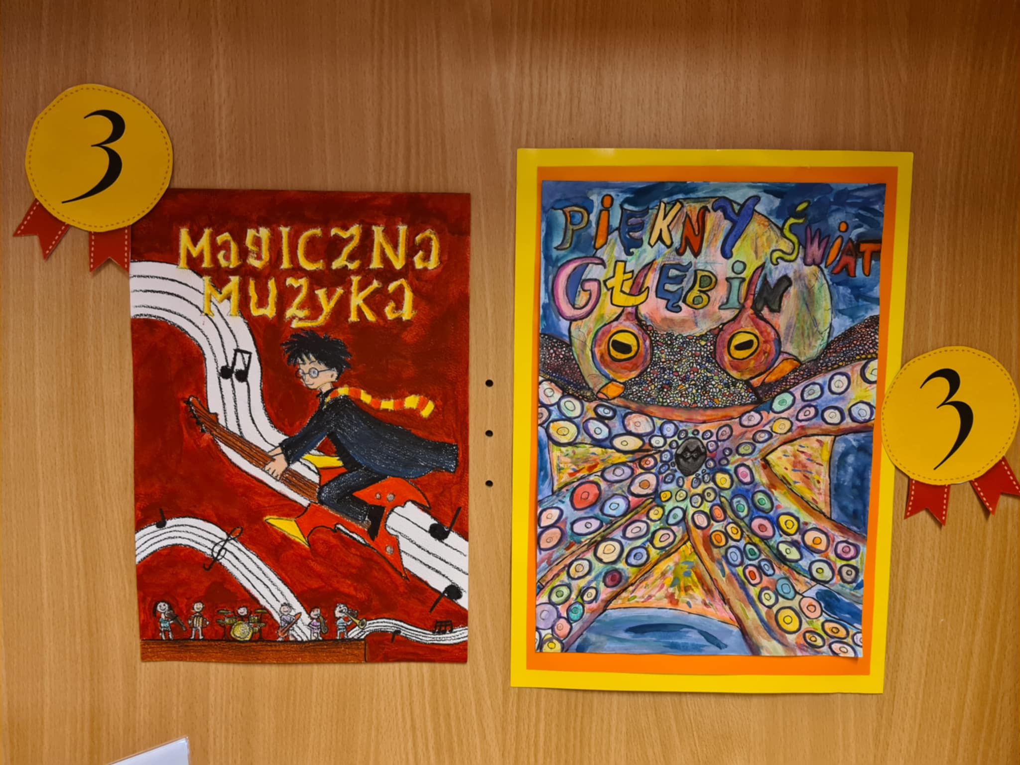Zbliżenie na dolny fragment półki z dwiema ilustracjami ex aequo na trzecim miejscu. Po lewej „Magiczna muzyka” — chłopiec w pelerynie grający na gitarze nad zespołem muzyków. Po prawej „Piękny świat głębin” — kolorowy ośmiornica z dużymi oczami i wieloma wzorzystymi mackami.