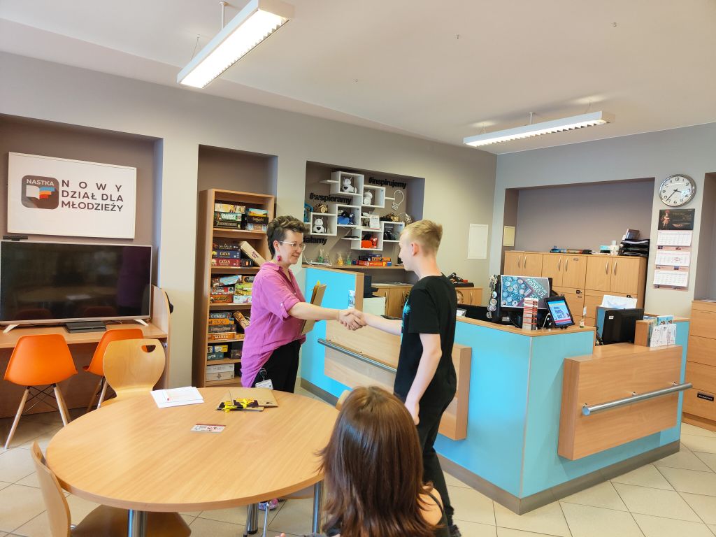 Na zdjęciu widać ceremonię wręczania nagród podczas konkursu plastycznego w bibliotece. W centralnej części kadru kobieta trzyma dyplom i ściska rękę jednemu z nagrodzonych chłopców. W tle widoczna jest młodzież siedząca przy stołach.