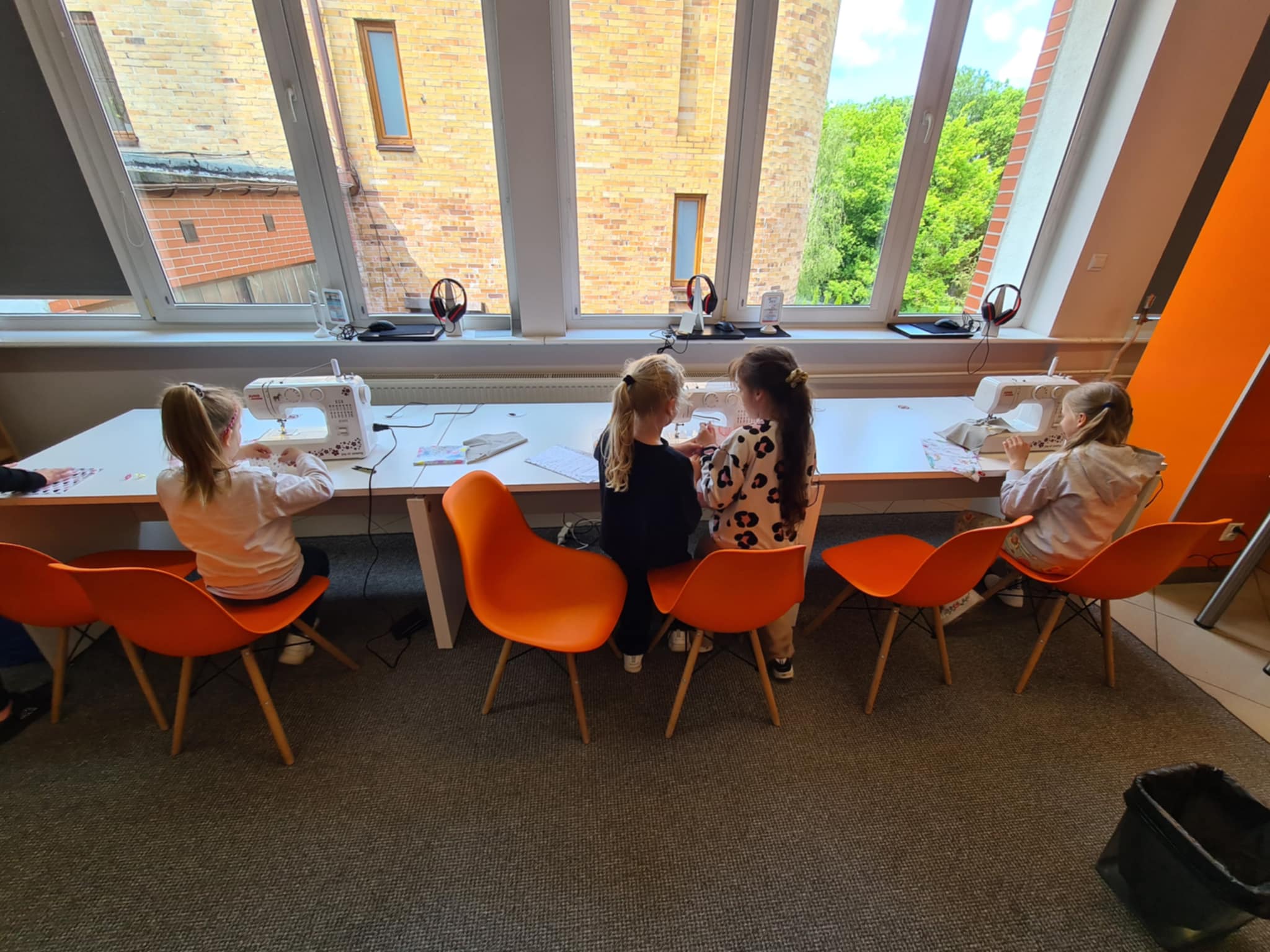 Kilkoro dzieci siedzi przy białych stołach i szyje na maszynach do szycia podczas warsztatów w bibliotece.