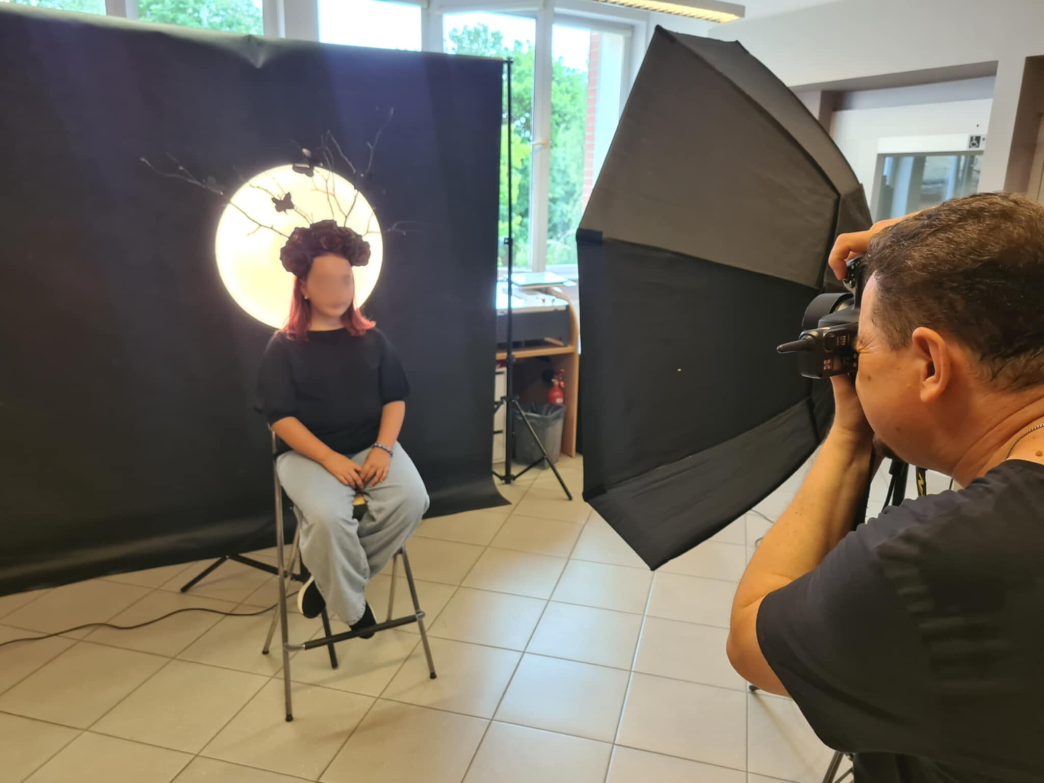 Modelka w czarnym nakryciu głowy stylizowanym na gałęzie i róże, fotograf przygotowuje ujęcie.