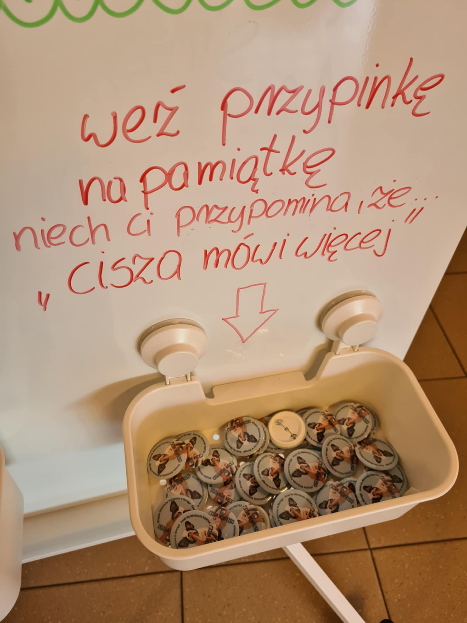 Pojemnik z pamiątkowymi przypinkami z grafiką psa i hasłem ‘cisza mówi więcej’.