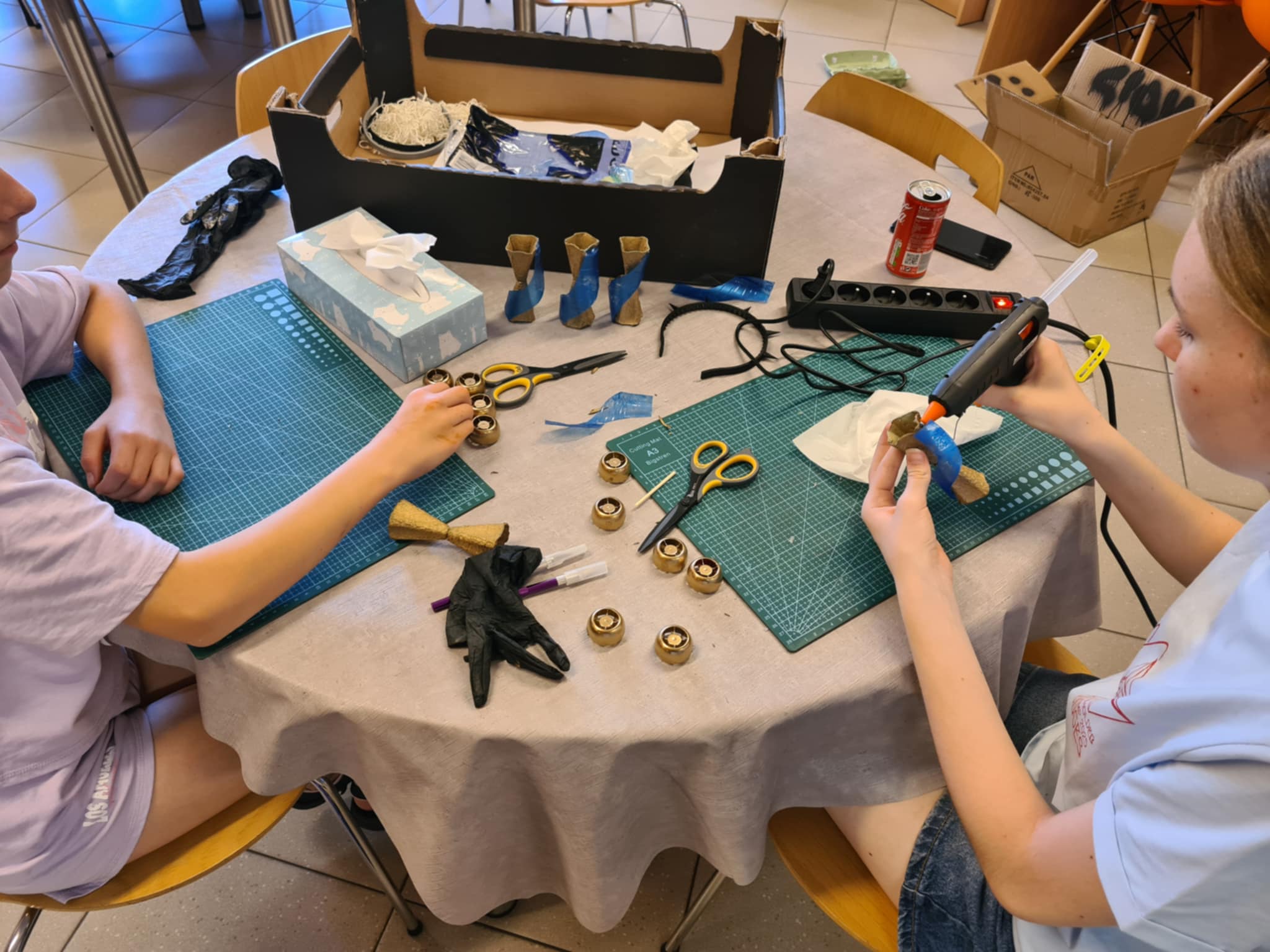 Dwie dziewczynki przy stole tworzą ozdoby z recyklingu, jedna używa pistoletu do kleju.