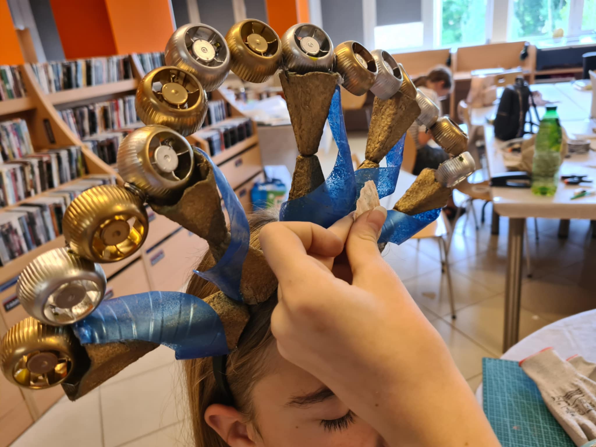 Dziewczynka zakłada ozdobną koronę z recyklingu z niebieskimi wstążkami i złotymi elementami.