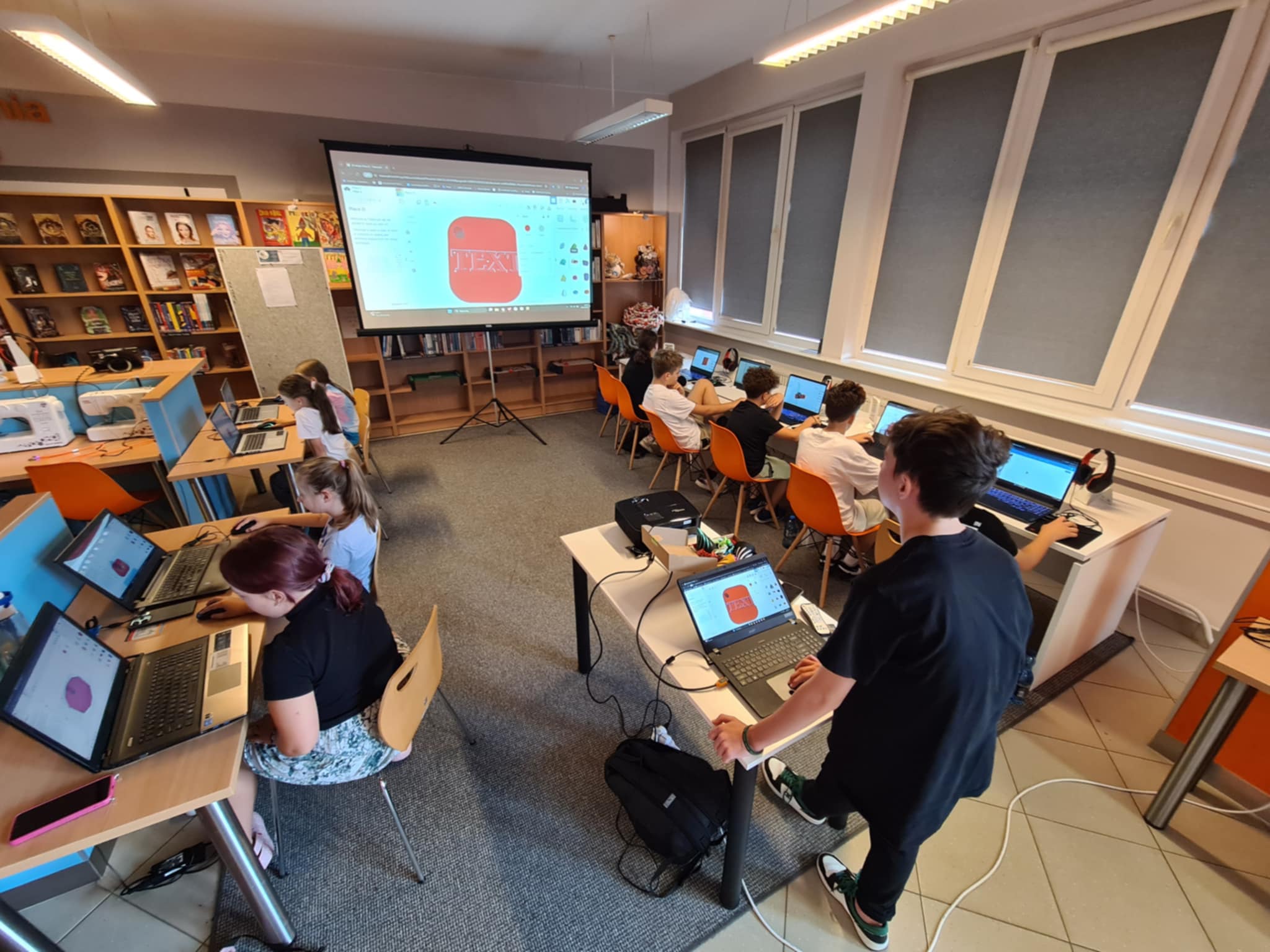 Widok sali warsztatowej w bibliotece, dzieci projektują obiekty 3D na laptopach, a na dużym ekranie widać czerwony model z napisem 'TEXT'.