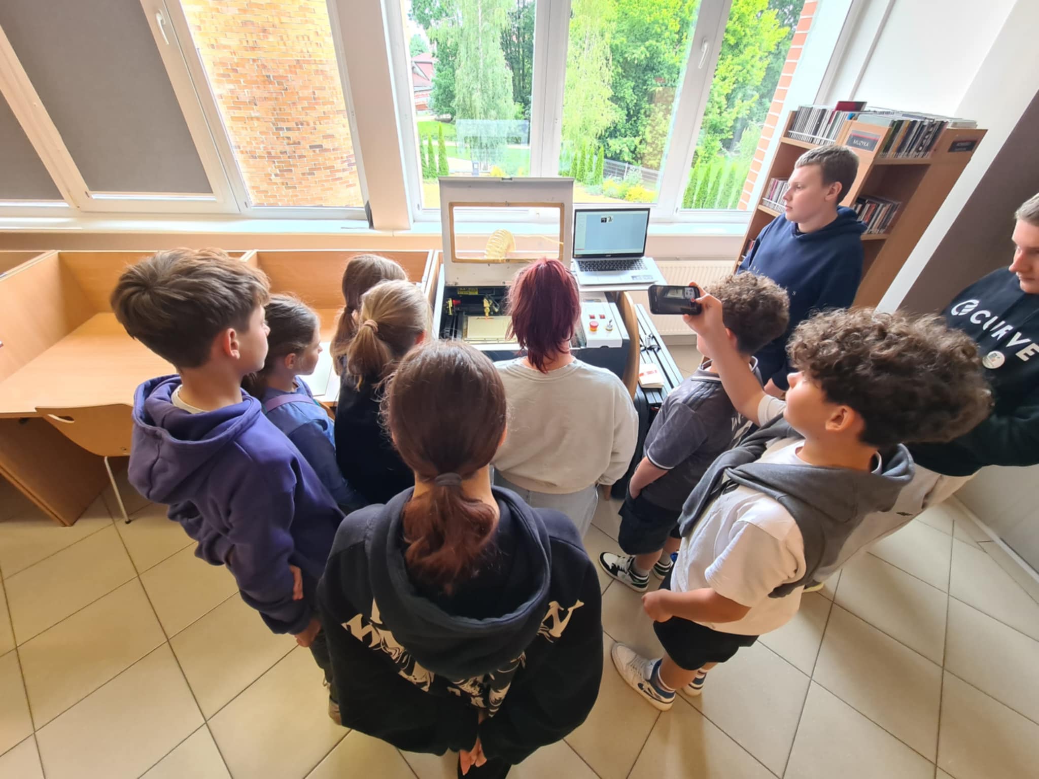 Zaciekawiona młodzież obserwuje działanie plotera laserowego podczas grawerowania.