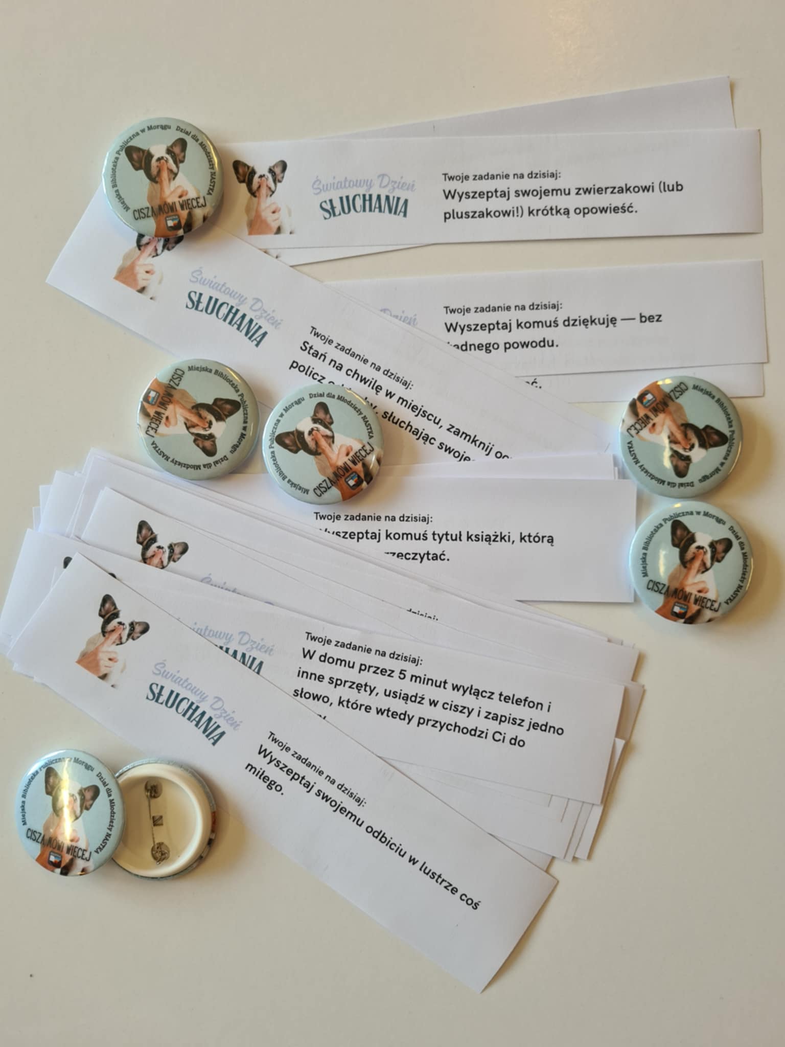 Zbliżenie na przypinki z grafiką psa i hasłem ‘Cisza mówi więcej’ oraz papierowe paski z zadaniami na Światowy Dzień Słuchania. Przykładowe zadania to: ‘Wyszeptaj swojemu zwierzakowi opowieść’, ‘Wyszeptaj komuś tytuł książki, którą chcesz przeczytać’, ‘W domu przez 5 minut wyłącz telefon i zapisz jedno słowo, które przychodzi ci do głowy’, ‘Wyszeptaj sobie coś miłego do lustra’.
