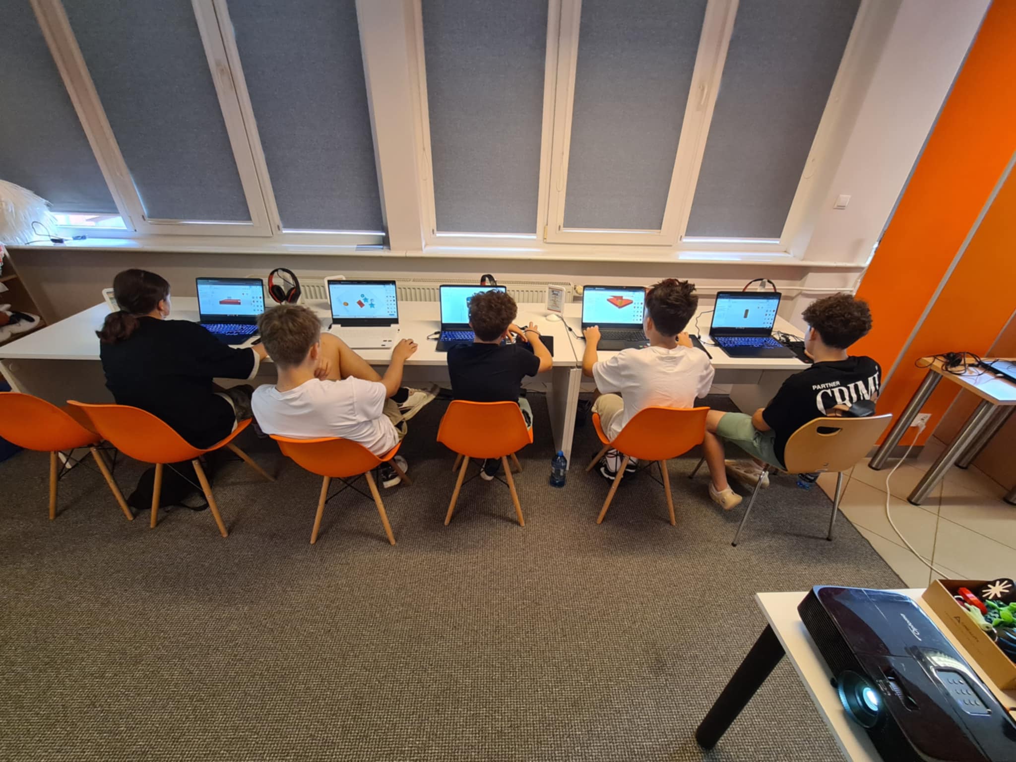 Grupa młodzieży projektuje modele 3D przy komputerach, siedząc ramię w ramię w nowoczesnej przestrzeni bibliotecznej.