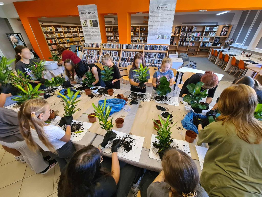 Grupa młodzieży podczas warsztatów w bibliotece – uczestnicy siedzą wokół stołu, w rękach trzymają rośliny przygotowane do techniki Kokedama.