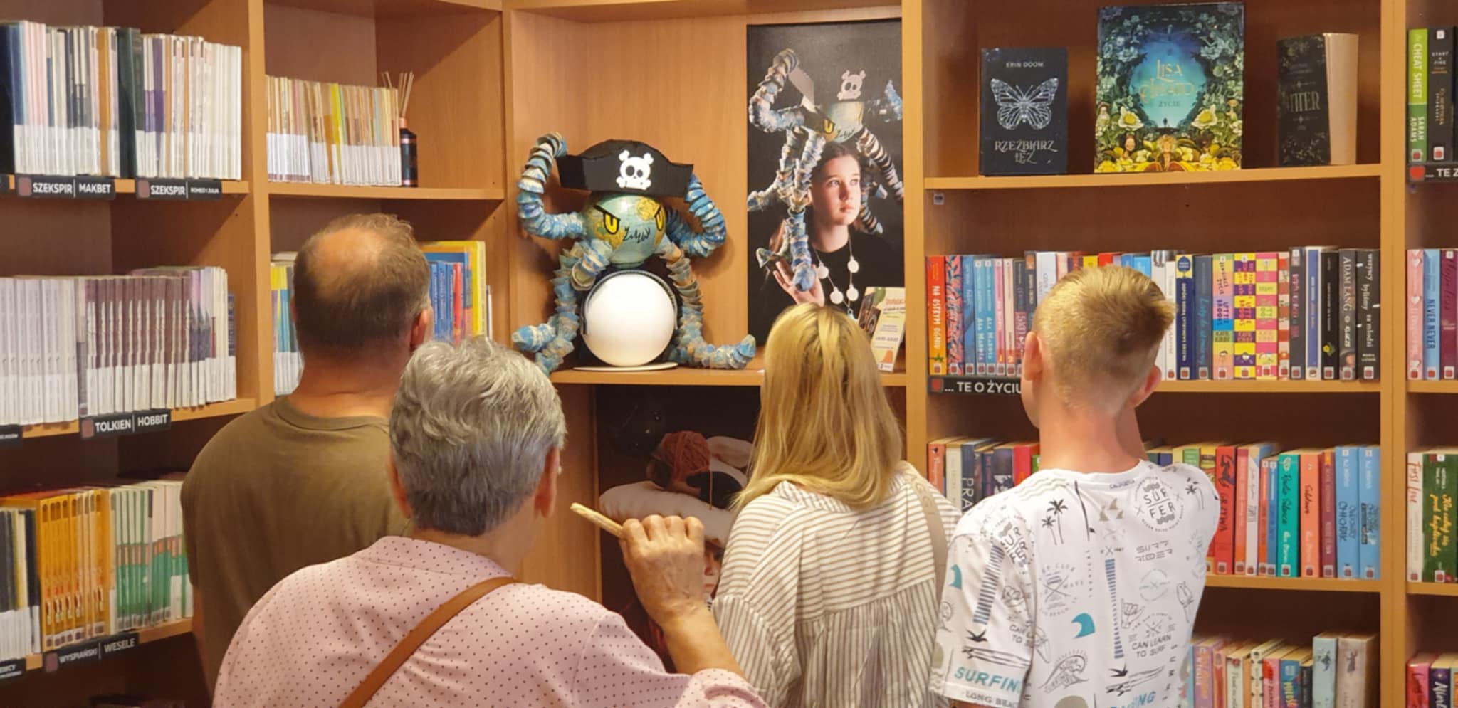 Zwiedzający oglądają pracę z wystawy EkoKreacje – grupa osób stoi przed półką z książkami, na której znajduje się rzeźba ośmiornicy w czapce pirata oraz zdjęcie modelki w nakryciu głowy inspirowanym tym motywem.