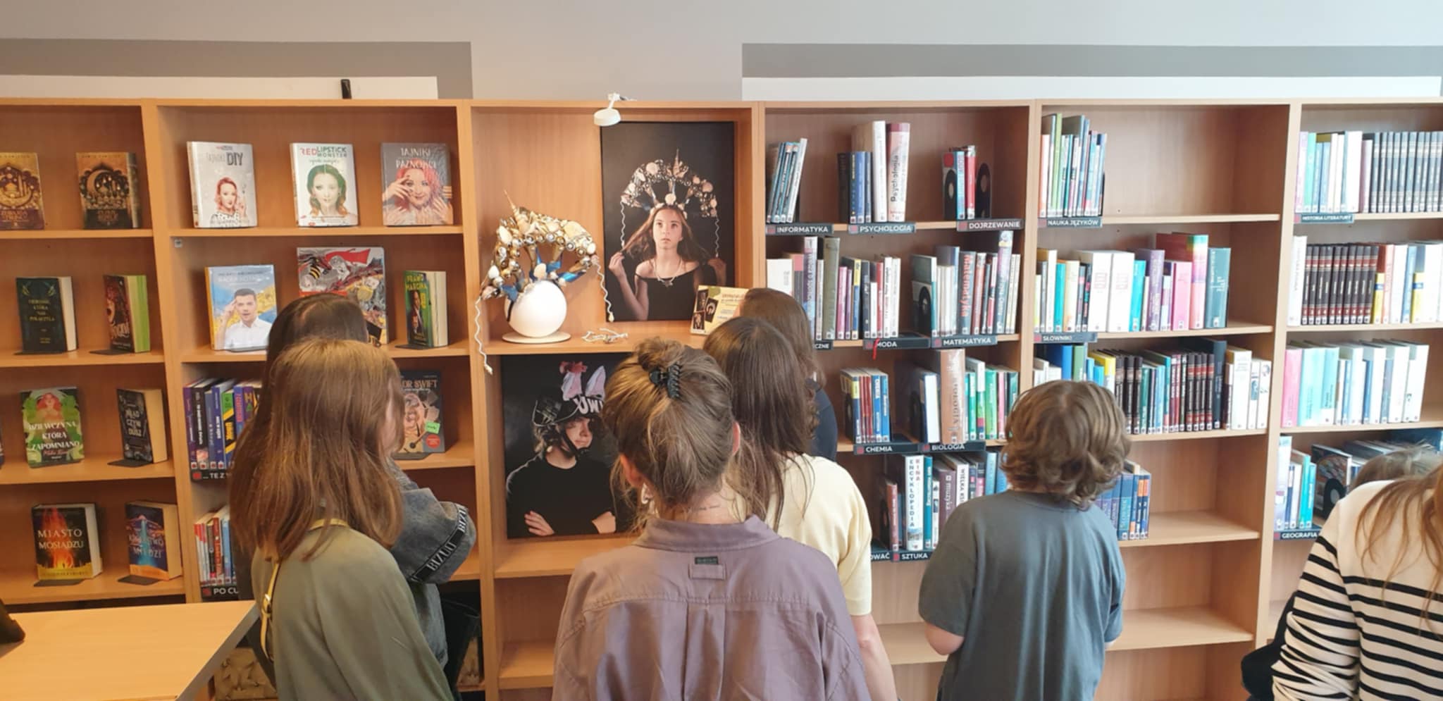 Grupa młodzieży ogląda w bibliotece wystawę kapeluszy z recyklingu i towarzyszących im fotografii.