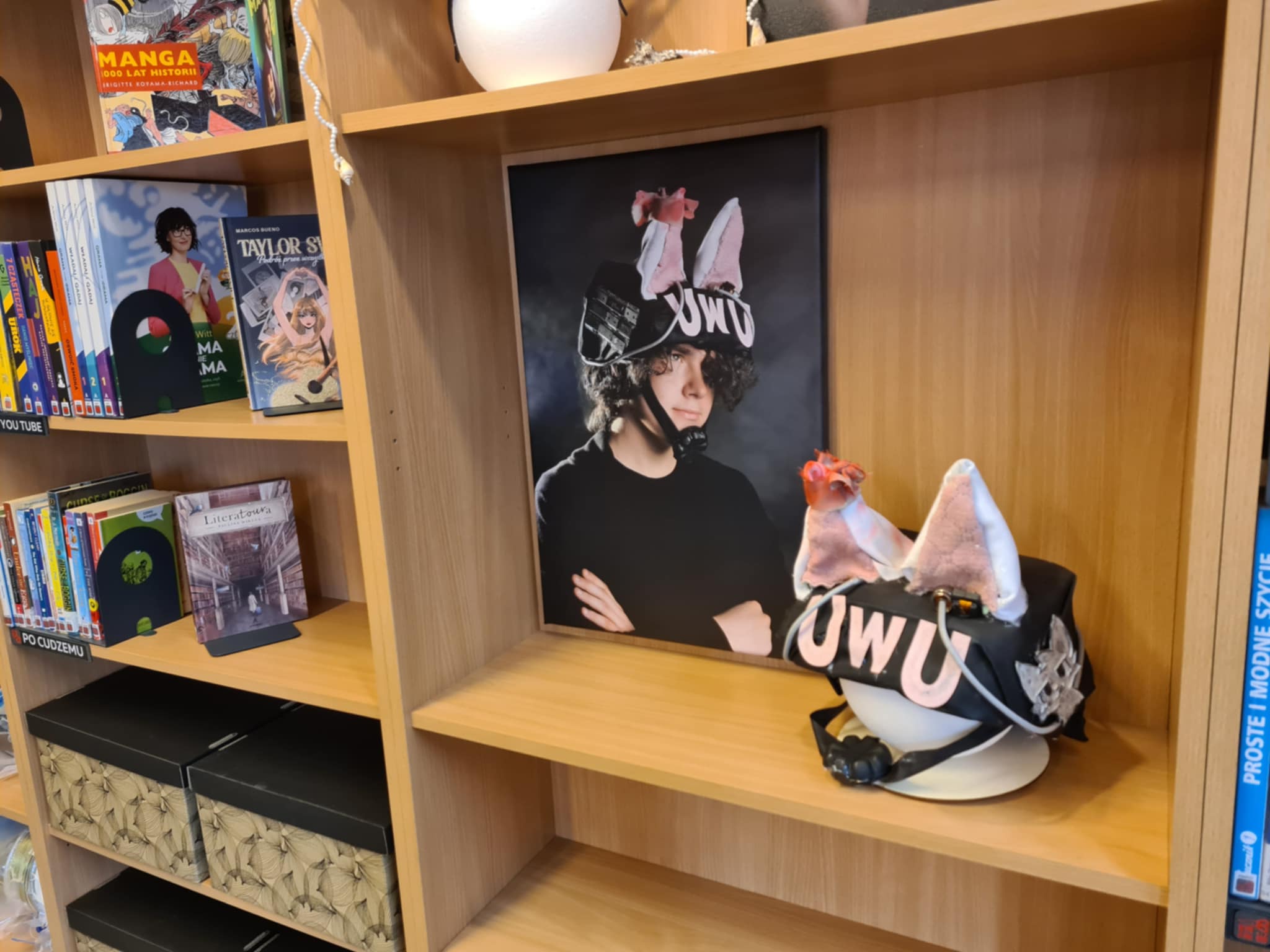 Półka biblioteczna z fotografią chłopaka w czarnym kapeluszu z uszami i napisem „UWU” oraz tym samym nakryciem głowy obok.