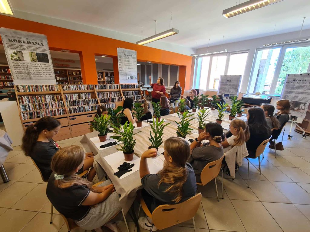 Warsztaty w pełni – uczestnicy siedzą wokół stołów w bibliotece, przed każdym z nich stoją małe doniczki i rośliny.