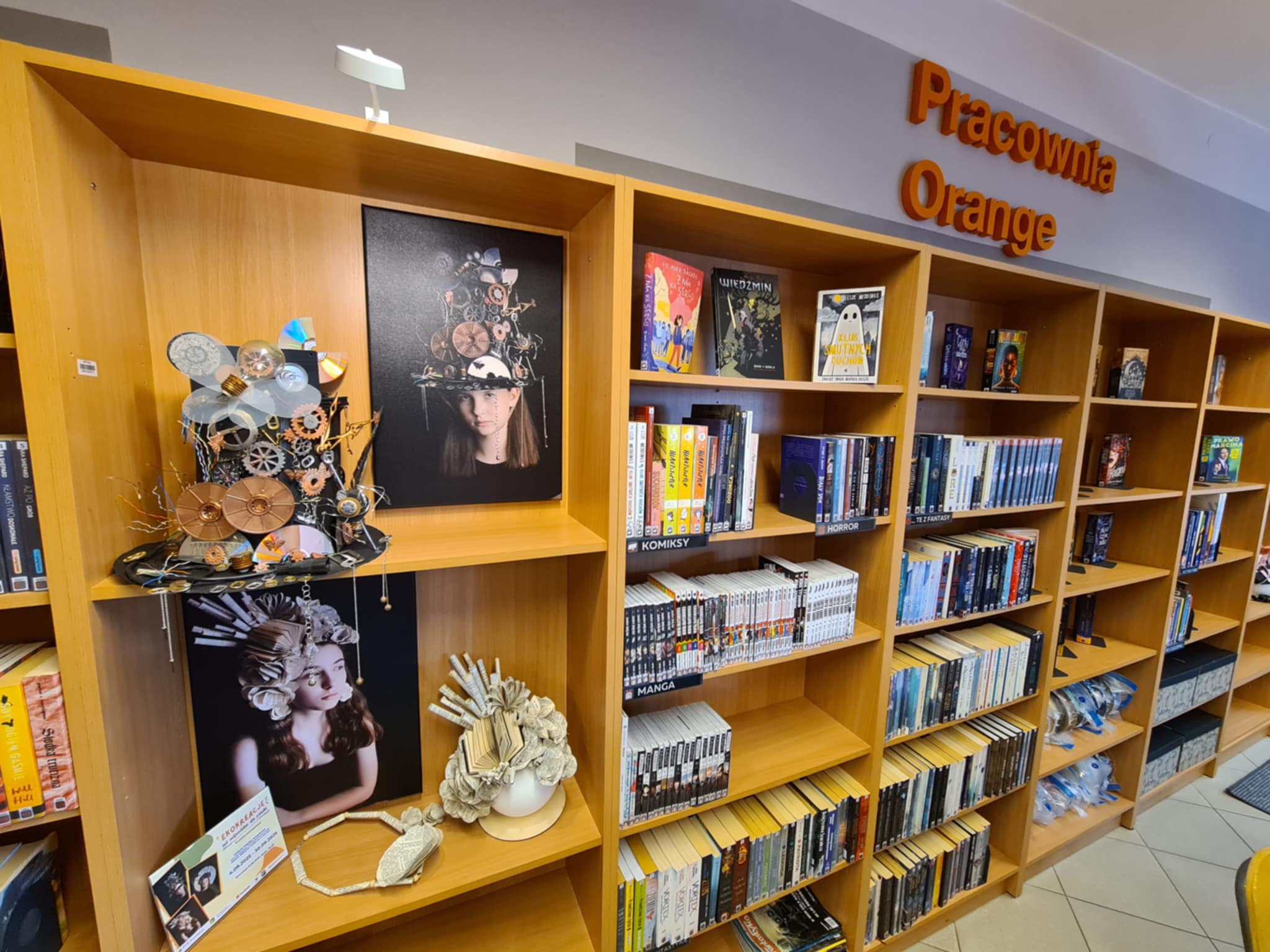 Wystawa w bibliotece z fotografiami dziewczyn w nakryciach głowy z recyklingu, w tle regał z książkami i napisem „Pracownia Orange”.