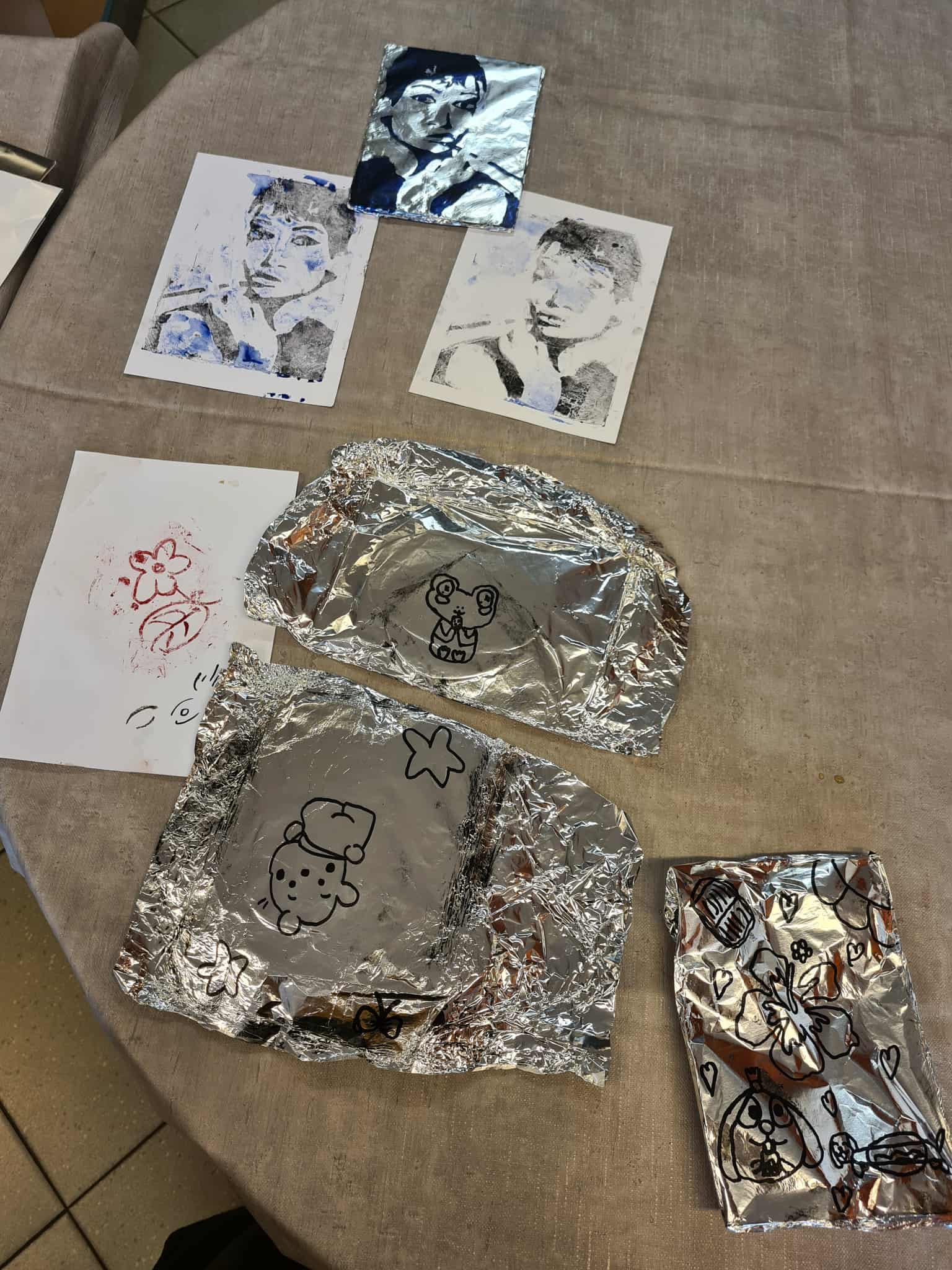 Efekty warsztatów: na stole leżą prace wykonane na folii aluminiowej i ich odbitki na papierze – portrety oraz proste rysunki.