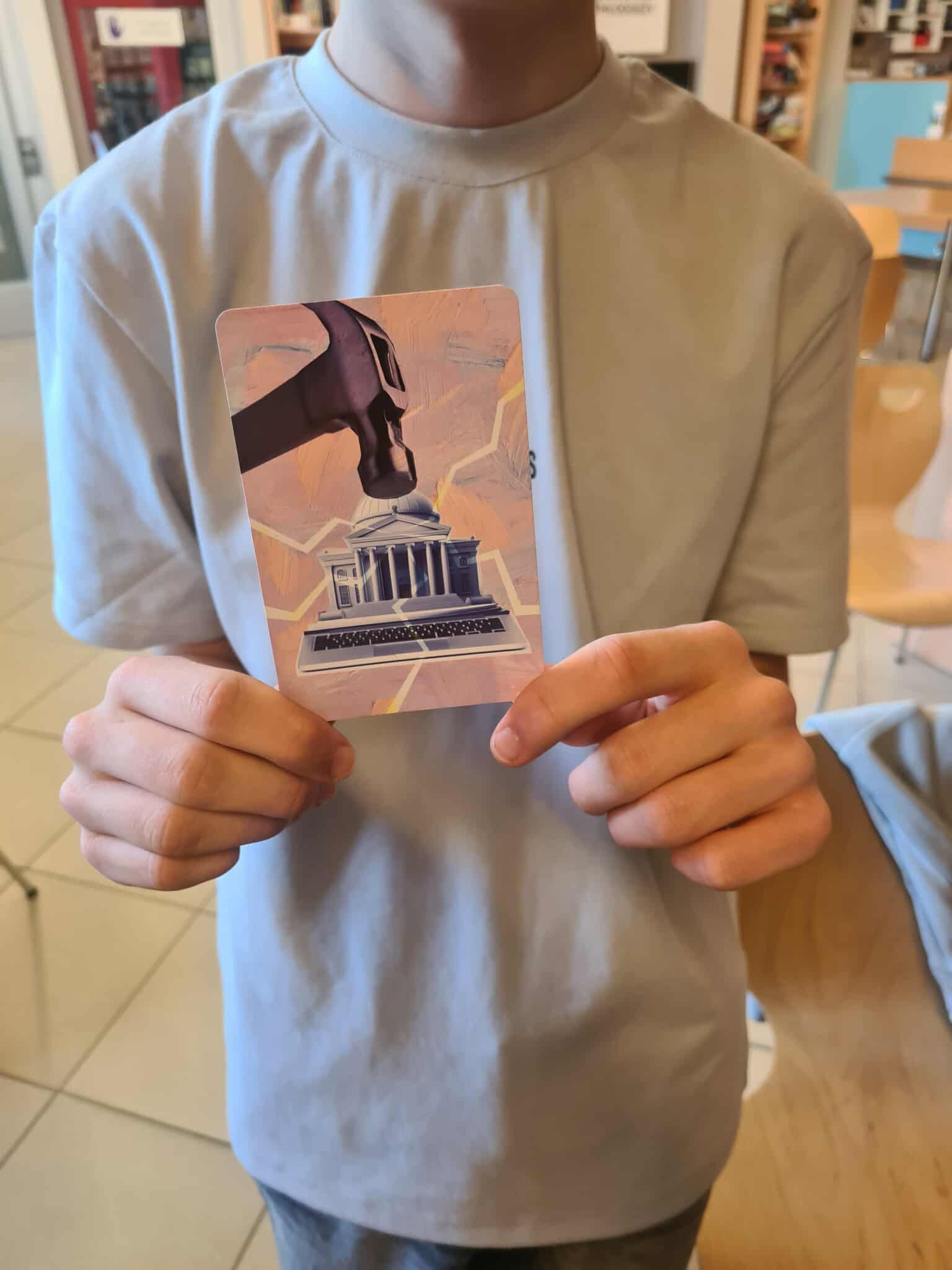 Osoba w jasnej koszulce trzyma kartę z ilustracją młotka nad budynkiem z kolumnami i klawiaturą laptopa.