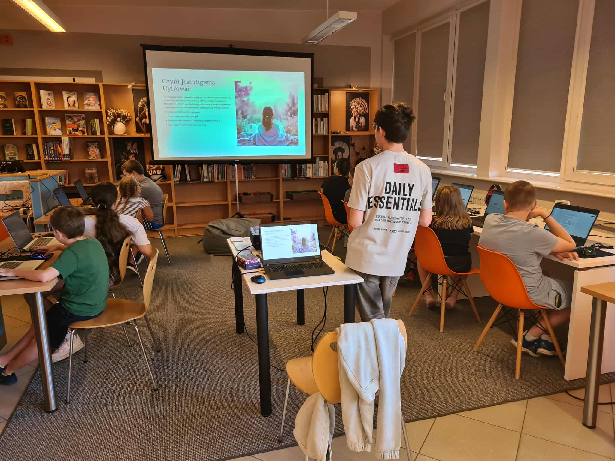 Grupa młodzieży w bibliotece pracuje na laptopach, oglądając prezentację o higienie cyfrowej.