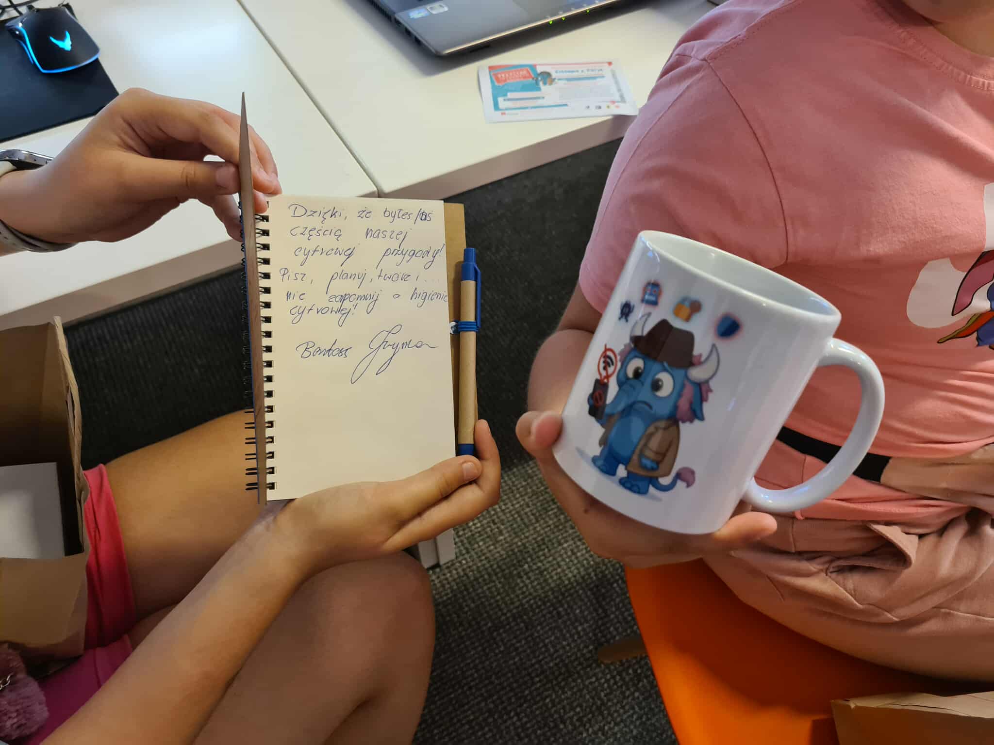 Osoba trzyma notes z odręcznym podziękowaniem i kubek z niebieskim słoniem w kapeluszu.