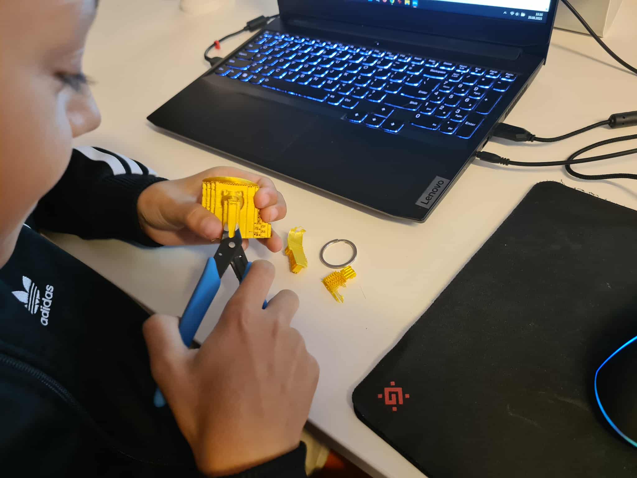 Chłopiec obrabia wydruk 3D – dziecko trzyma żółty wydruk 3D i używa małych szczypiec do usuwania podpór. Obok leży laptop Lenovo i czarny podkładka pod mysz.