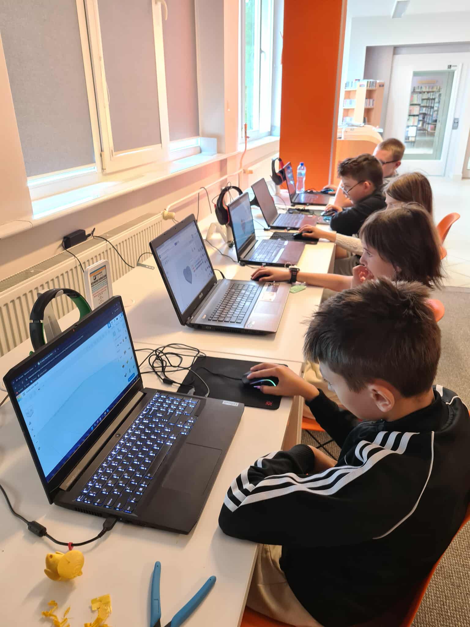Dzieci projektujące w Tinkercadzie – grupa dzieci siedzi przy komputerach w bibliotece, projektując modele 3D w programie Tinkercad.