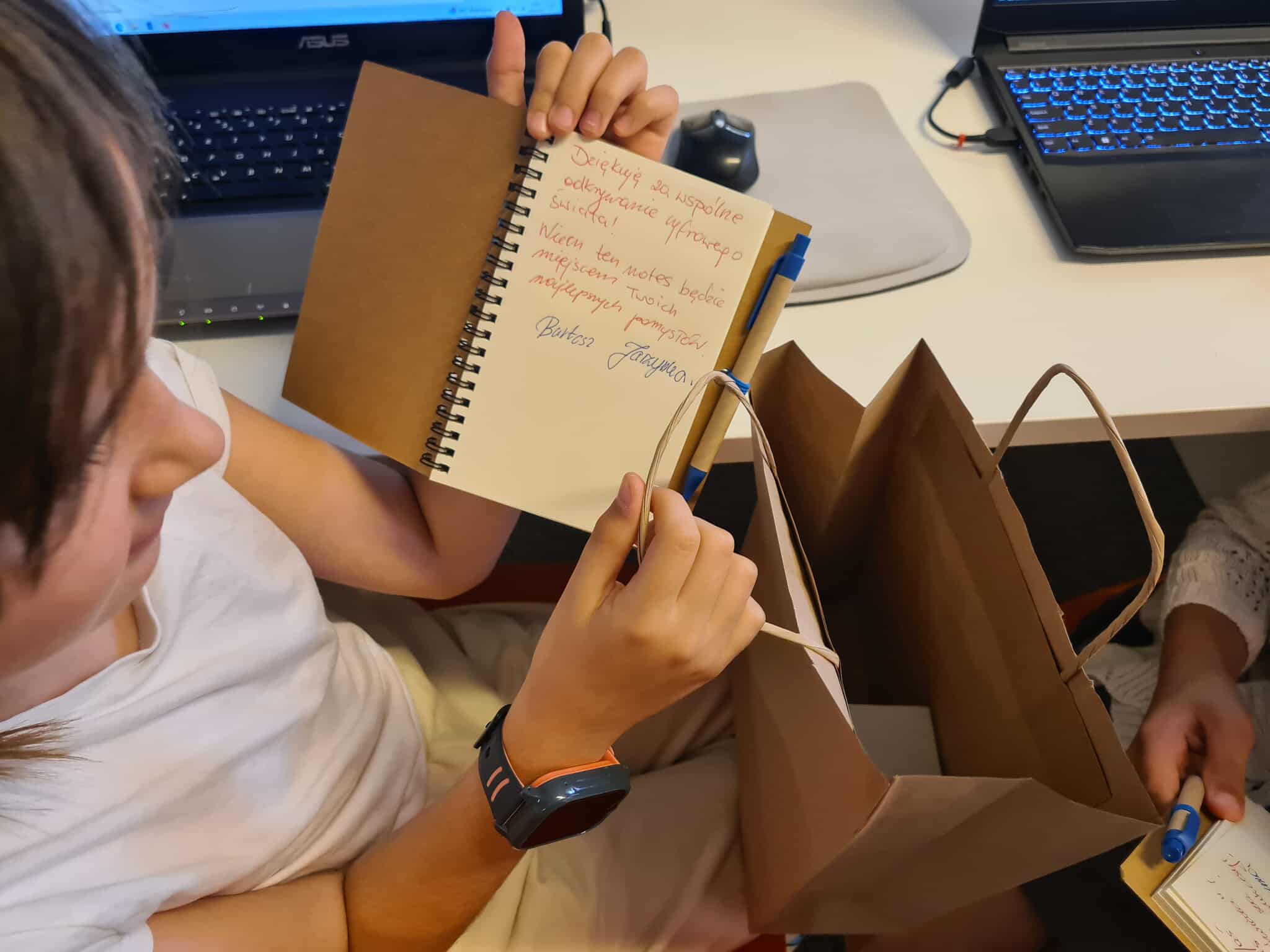 Notes z dedykacją – dziecko ogląda notes z ręcznie napisaną dedykacją i podpisem, w tle brązowa papierowa torba i długopis.