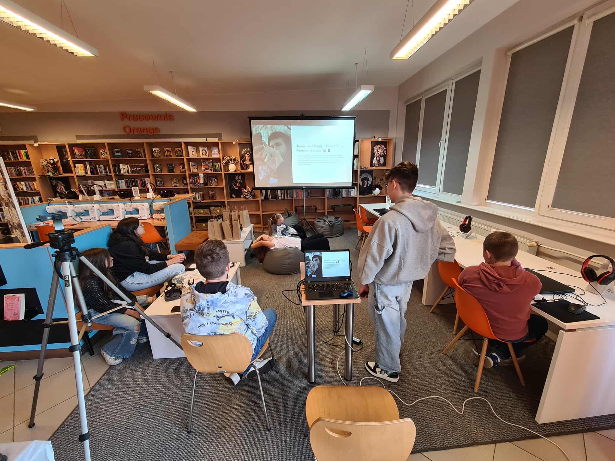 Grupa nastolatków podczas prezentacji w bibliotece, na ekranie widoczny slajd z tekstem i grafiką.