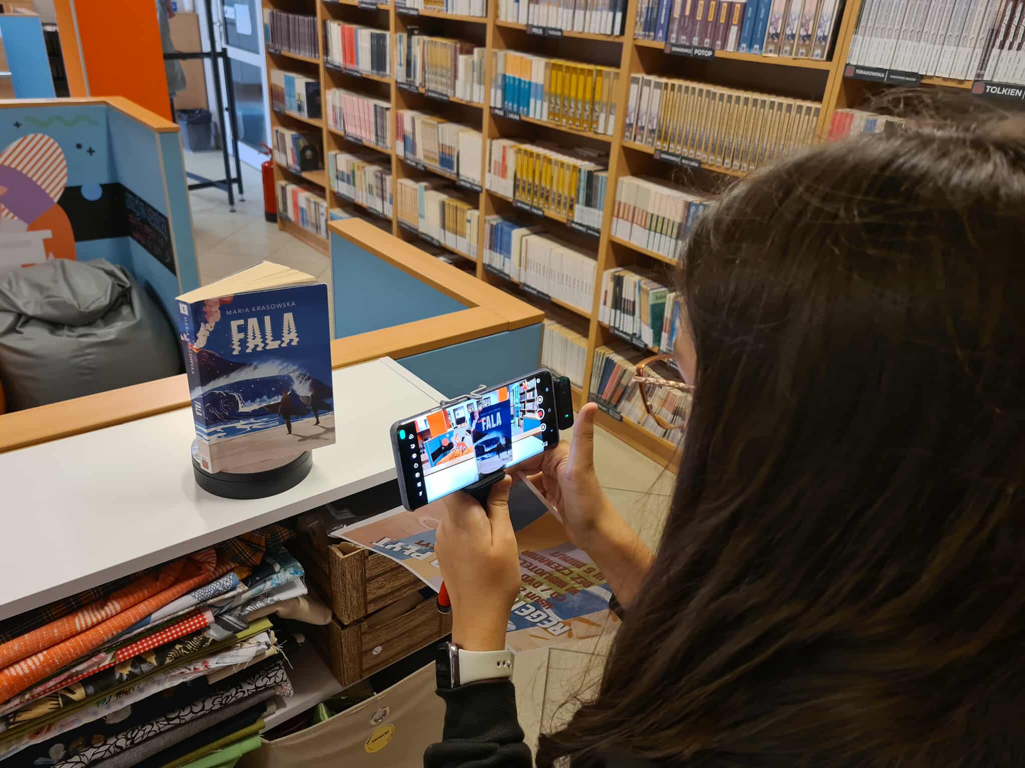 Dziewczyna fotografuje telefonem książkę „Fala” ustawioną na stojaku w bibliotece.