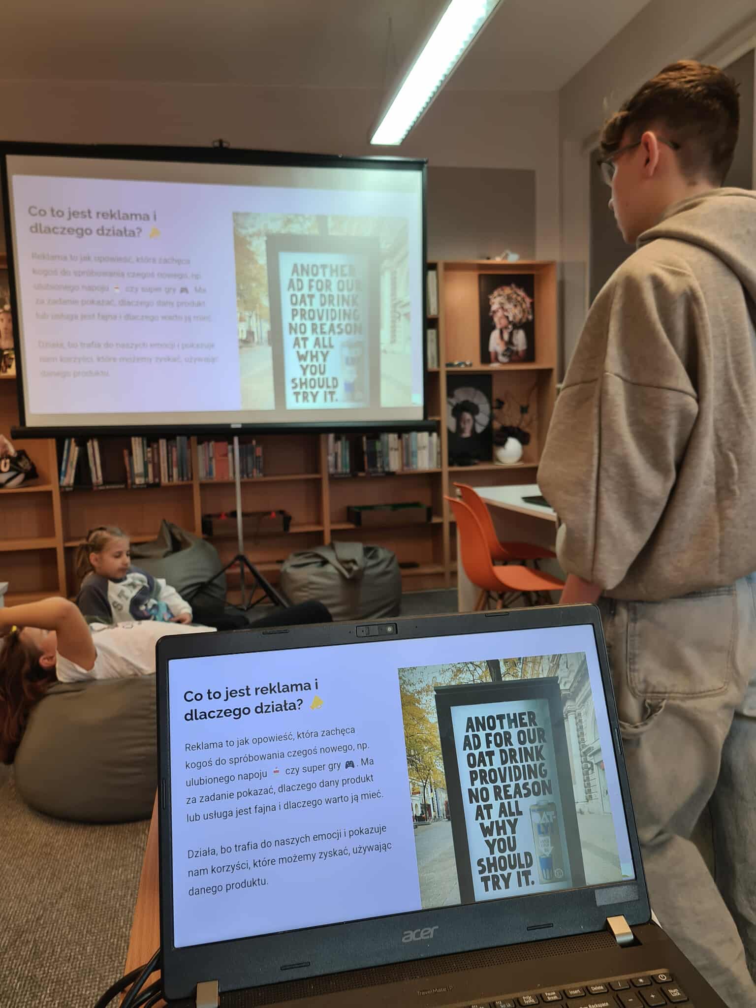 Chłopiec w szarej bluzie prezentuje slajd o reklamie na projektorze w bibliotece.
