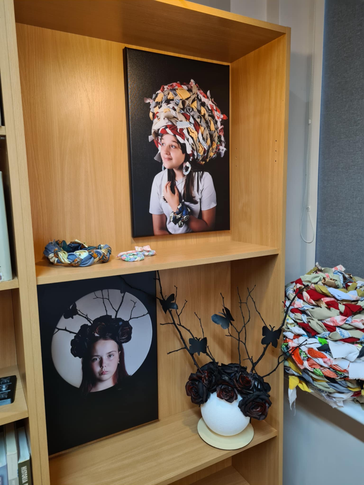 Półka biblioteczna z fotografią dziewczyny w dużym, kolorowym nakryciu głowy z materiału oraz czarną dekoracją z róż i motyli.