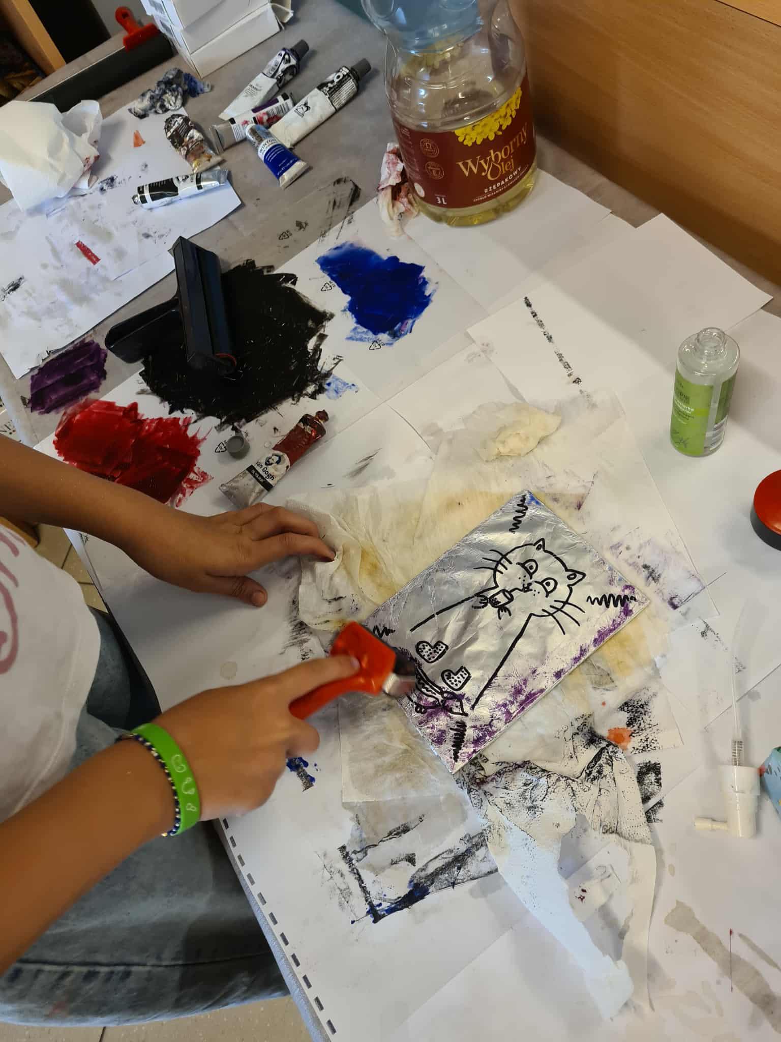 Uczestnik odbija rysunek kota z folii aluminiowej, używając małego wałka malarskiego. Na stole widać farby i butelkę oleju.