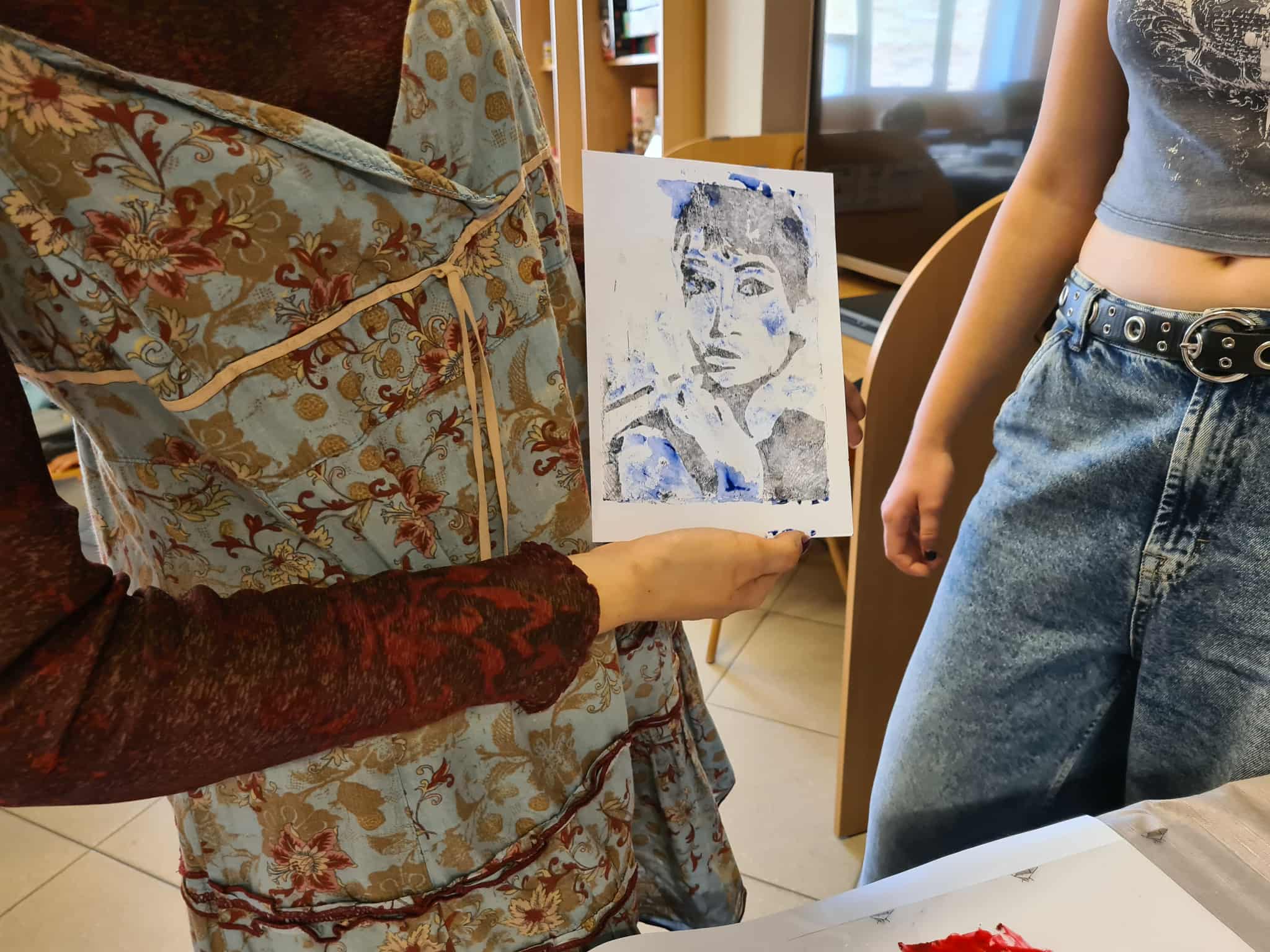 Instruktorka pokazuje odbitkę portretu zrobioną w technice graficznej. Obok niej stoi uczestniczka warsztatów.
