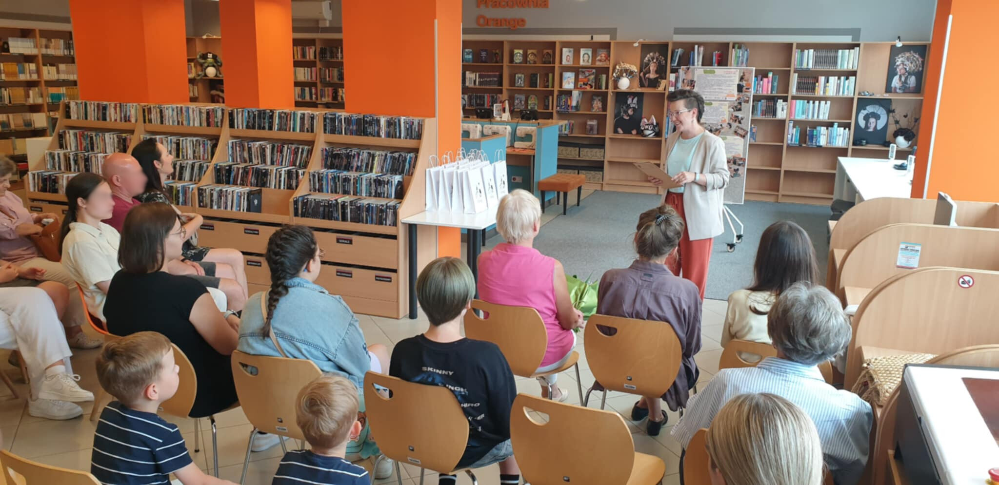 Grupa osób siedzi na krzesłach w bibliotece, przed nimi stoi kobieta w jasnych żakiecie