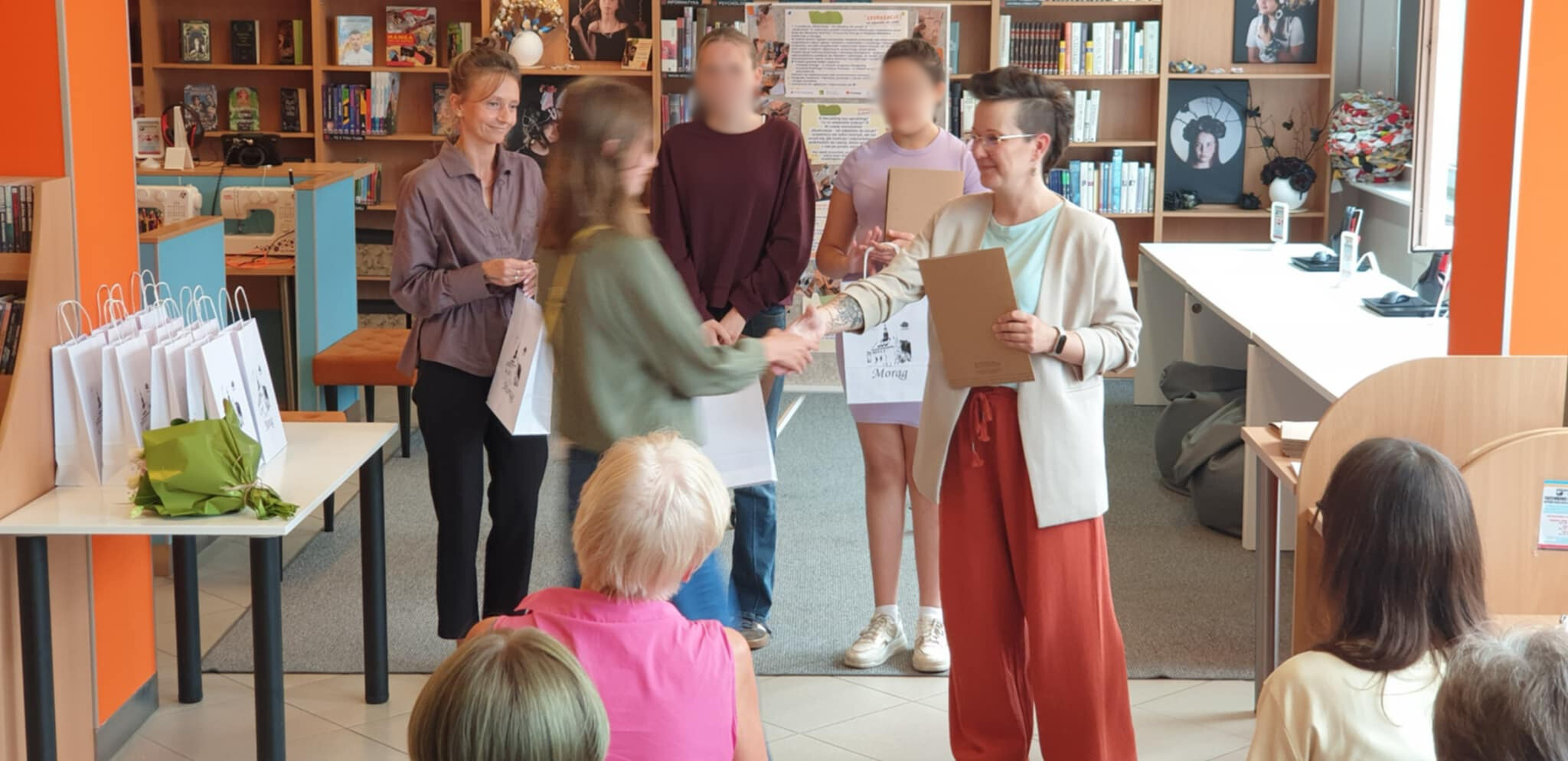 Wręczanie nagród uczestnikom projektu EkoKreacje w bibliotece – kobieta z krótkimi włosami i w jasnym żakiecie podaje dyplom młodej uczestniczce, w tle inne osoby z torbami upominkowymi.