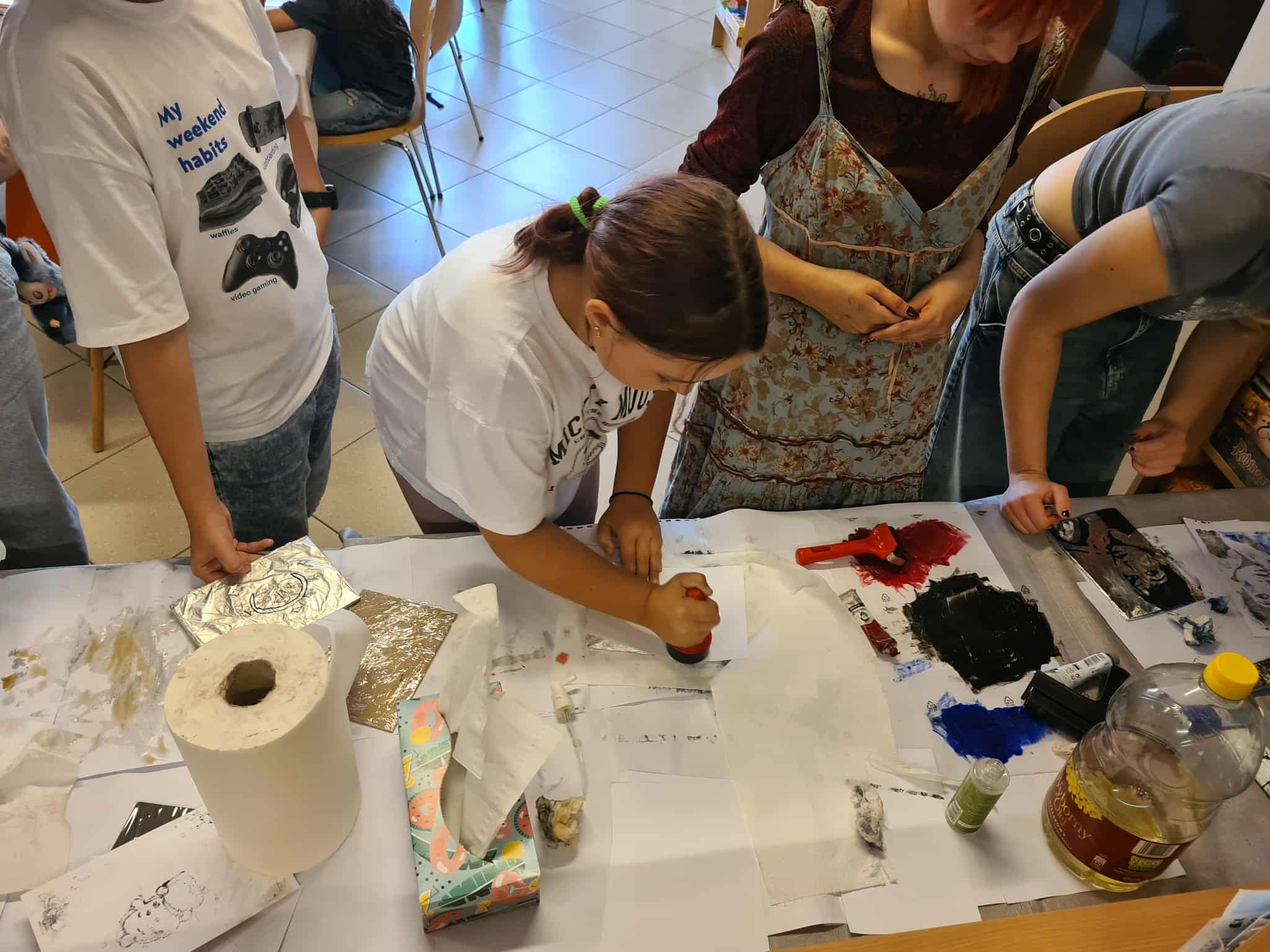 Grupa młodzieży pracuje przy stole, wykonując odbitki graficzne z użyciem farb, folii i prostych narzędzi. Na stole widać kolorowe farby i przybory plastyczne.