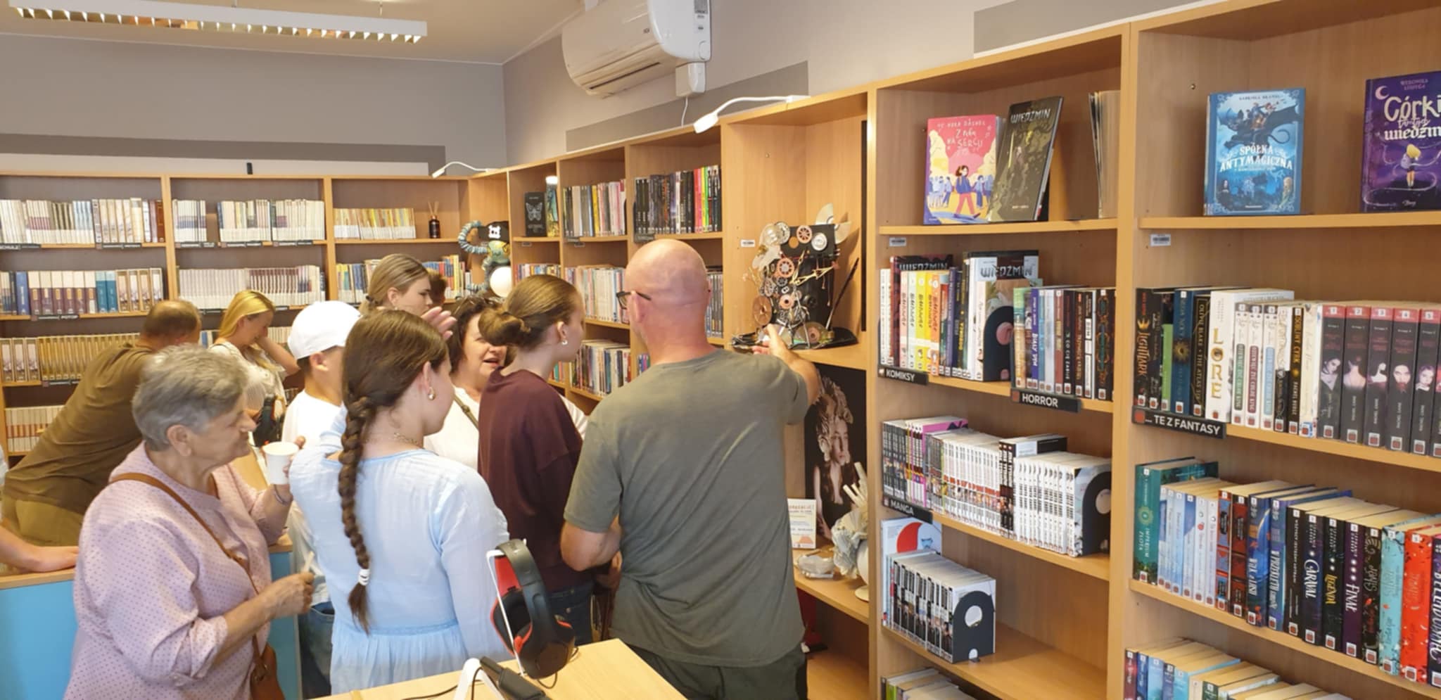 Zwiedzający podziwiają steampunkowy kapelusz na wystawie EkoKreacje – na półce stoi nakrycie głowy z elementami kół zębatych i miedzianych ozdób, obok portret modelki w tym kapeluszu, w tle regały z książkami.